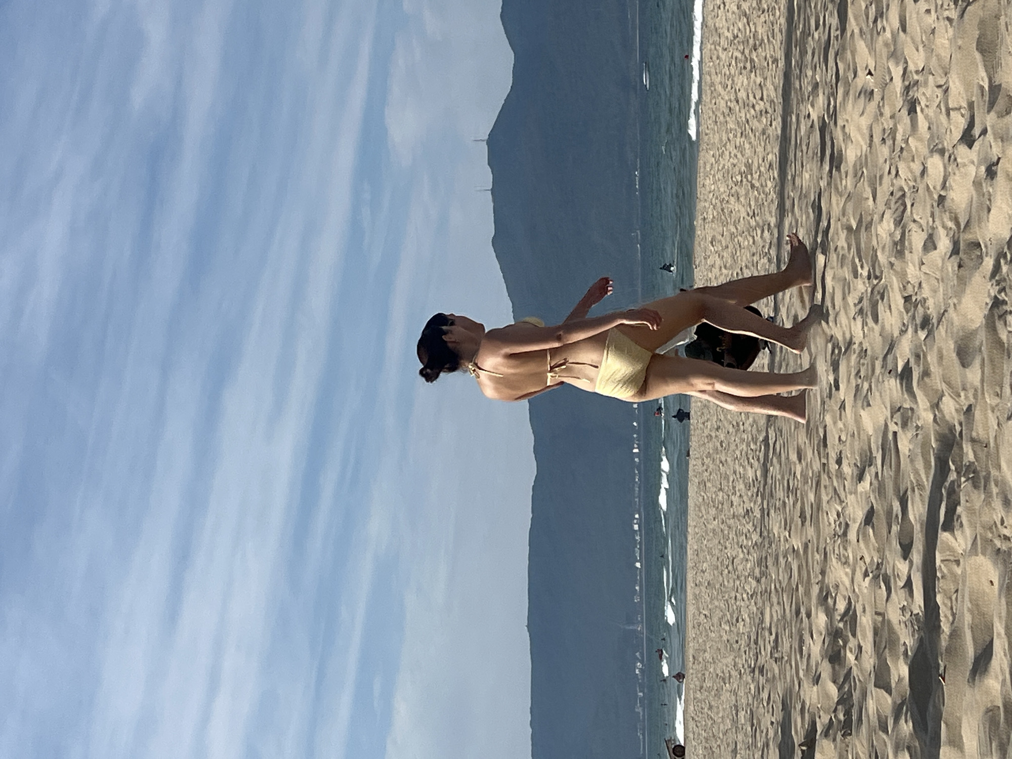 A woman in a yellow bikini walks on a sandy beach in Đà Nẵng, Vietnam, with the ocean and mountains in the background. The sky is partly cloudy with light blue streaks of clouds. The ocean has moderate waves, and there are a few people in the water and on the beach. The mountains in the distance are dark green and covered in trees, with some buildings visible at their base. The sand is light brown and textured, with shadows from ripples.