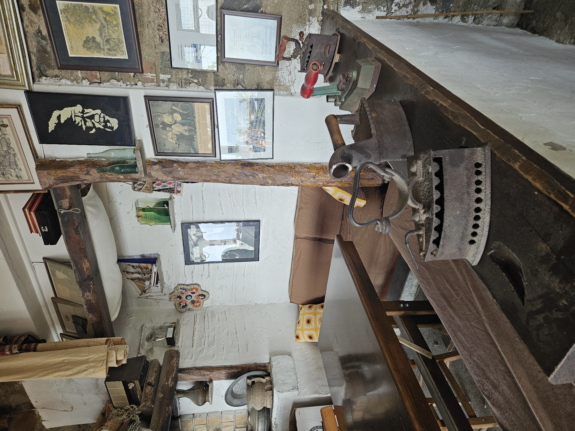 This interior image depicts a cultural or historical establishment in Tire, Turkey. In the immediate foreground, a dark wooden countertop displays a collection of five antique clothes irons of varying designs, including cast-iron models with wooden handles and one with a red handle, all exhibiting signs of rust and age. To the left, a polished dark wooden dining table with a visible chair leg occupies the foreground.
The midground features a brown upholstered bench or sofa positioned against the textured, white-washed walls. A yellow crocheted cushion with sunflower motifs rests on the sofa. The walls are adorned with numerous framed artworks and photographs. Notable items include a black framed artwork with a gold profile of a man's face, a framed black and white photograph captioned "Tire Bandosu 'Amerika yolcusu'" depicting a group of men with musical instruments, and a framed landscape drawing dated "Tire - 1984." Other framed images include portraits, a group photo, and a document. A small wooden sailing ship model hangs from a shelf, and two green glass bottles are placed on another small wooden shelf. A decorative evil eye wall hanging is visible on the wall.
To the far left, a white-washed built-in structure, possibly a fireplace or oven, serves as a display area for a dark pottery vase, a decorative metal plate, and a wooden mortar and pestle. Above this, a vintage radio receiver sits on a wooden shelf next to a woven basket and draped beige fabric. Exposed dark wooden beams and a vertical wooden post are prominent architectural features, contrasting with the rough, white-washed plaster walls. The floor is dark and indistinct. The scene is illuminated by ambient light, suggesting an indoor setting with a rustic, historical ambiance.