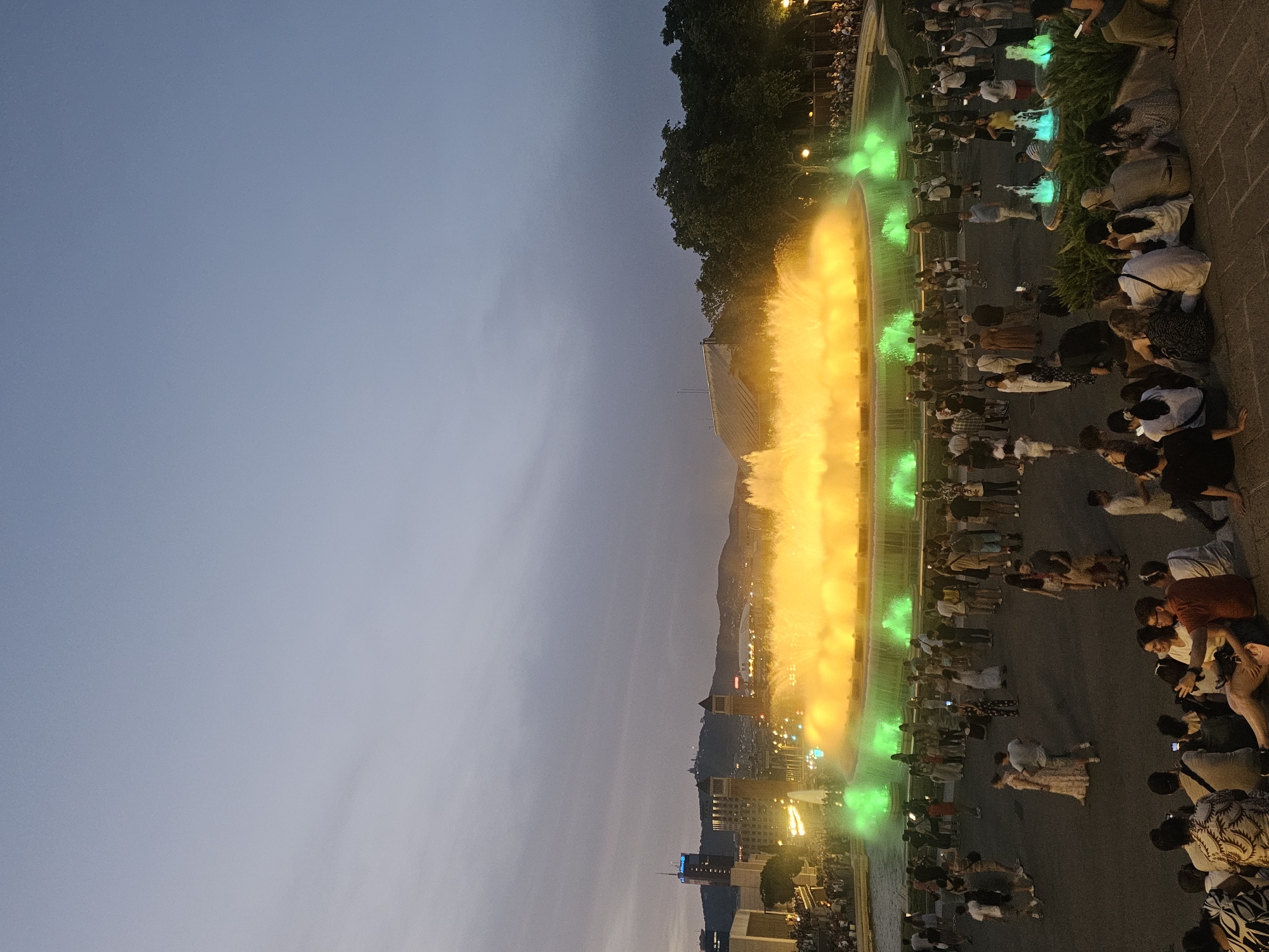 This is a daytime photograph of the Magic Fountain of Montjuïc in Barcelona, Spain. The fountain is illuminated with vibrant yellow and green lights, casting a warm glow on the surrounding area. A large crowd of people are gathered around the fountain, enjoying the spectacle. Many are sitting on the steps leading up to the fountain, while others stand and watch from a distance. In the background, the city skyline is visible, with several buildings and a hill in the distance. The sky is a soft blue with some clouds. The scene appears to be a popular tourist attraction, with people of all ages enjoying the beautiful display of light and water.
