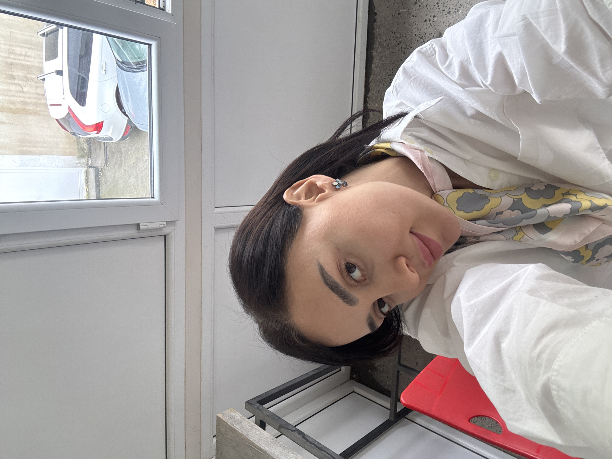 A woman with dark hair is visible from the shoulders up, wearing a white shirt and a patterned scarf. She is positioned at an angle, with a white window and an exterior view visible to the left. The exterior reveals a car and the side of a building or street, suggesting an urban setting. The background also includes a white door or wall panel, and a portion of what appears to be a chair or stool with a red seat. The specific location is not discernible from the image alone.