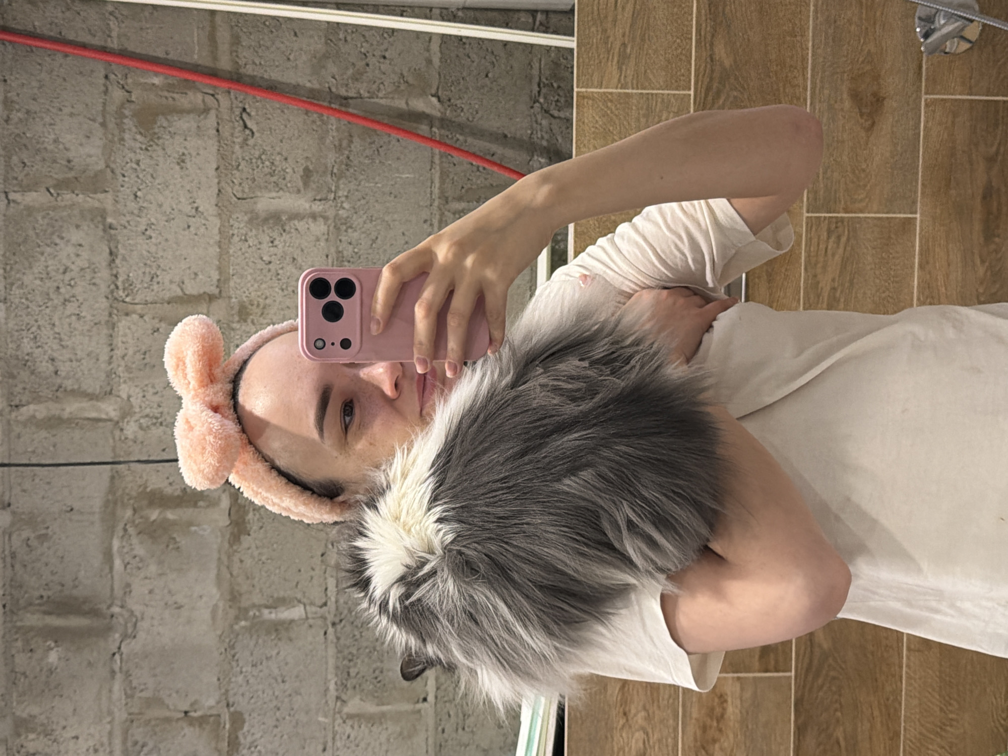 A person with a pink bunny ear headband is holding a gray and white cat while taking a selfie with a pink phone. The background includes a brick wall and tiled wall with a shower head. The location is Chaikovsky, Russia.