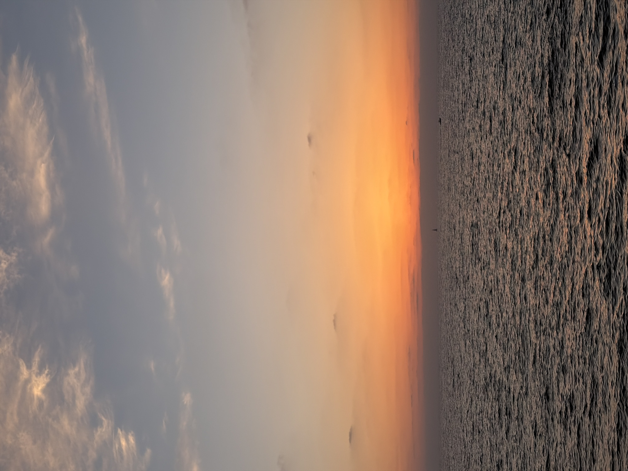 The image shows a calm sea at sunset, viewed from the coast of Tel Aviv, Israel. The sky is a gradient of colors, from pale blue with wispy clouds at the top to vibrant orange and pink near the horizon. The sea is dark and choppy, reflecting the colors of the sky. In the distance, two small boats can be seen on the water. The scene is serene and peaceful, evoking a sense of tranquility.