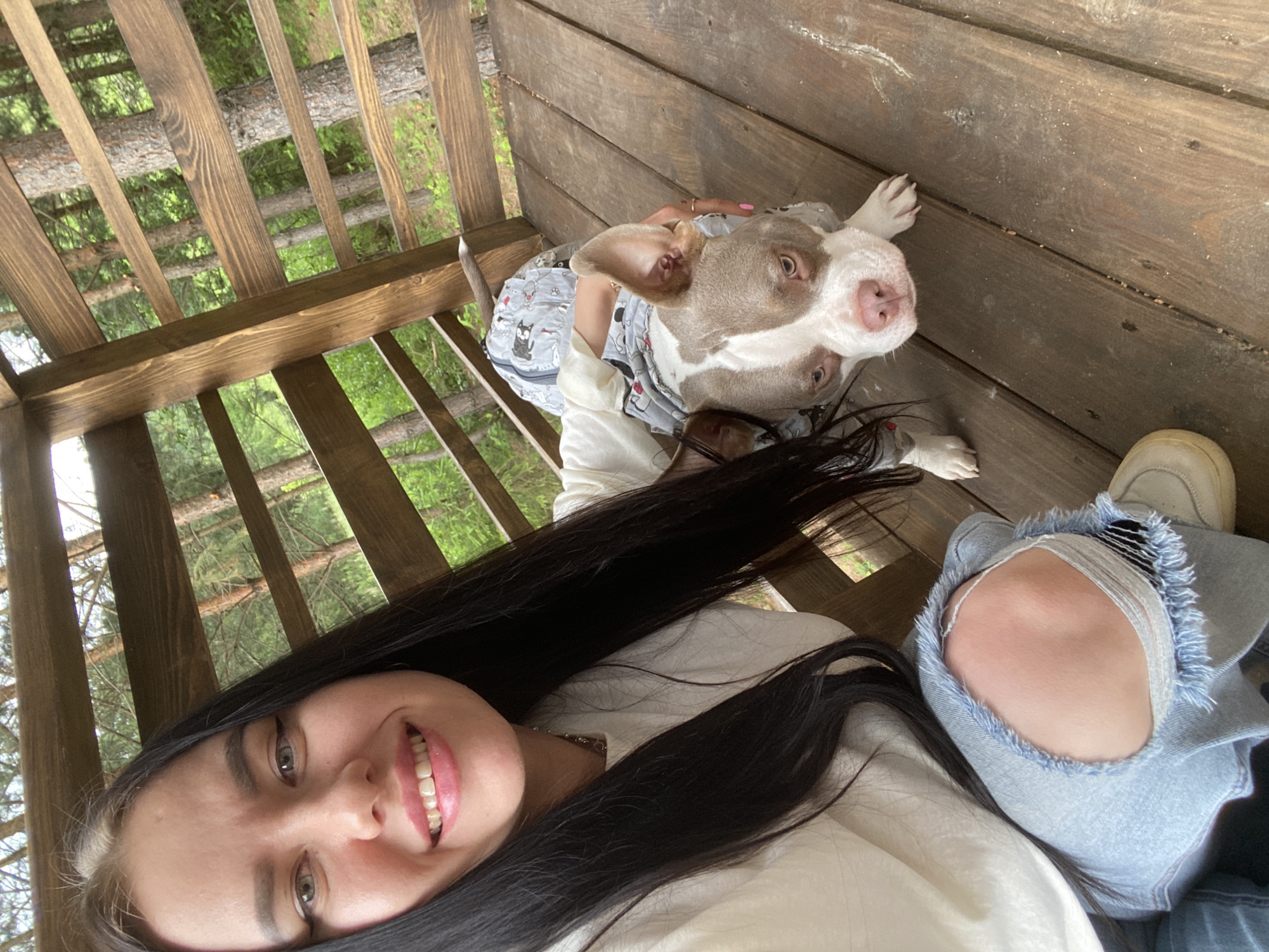 A young woman with long, dark hair and a bright smile is taking a selfie on a wooden deck surrounded by trees. She is wearing a white t-shirt and ripped denim jeans. Next to her is a dog, likely a pit bull, wearing a grey and white patterned outfit. The dog is looking directly at the camera. The background shows a forest with tall trees and wooden railings, suggesting an outdoor setting perhaps in a park or a residential area in the city of Fokino, Russia.