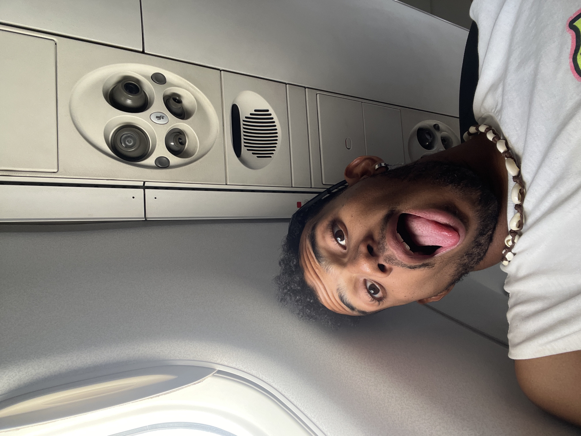 This media file is a close-up, low-angle selfie taken from inside an airplane cabin. The subject is a young man with curly dark hair, a beard, and a playful expression, sticking out his tongue towards the camera. He is wearing a white t-shirt and a necklace made of shells and beads.
The background of the image reveals typical airplane interior features. On the ceiling above the man, there is an overhead panel with what appears to be air vents and reading lights. To the left, a portion of an airplane window is visible, with natural light illuminating the cabin. The overall lighting suggests it is daytime.
The man's exaggerated facial expression and the candid nature of the selfie indicate a moment of fun or a lighthearted attempt to capture his travel experience. The orientation of the image, being taken from below and looking up, adds to the informal and playful tone. The location cue of Cumbo, Venezuela, implies the man is either departing from, arriving in, or in transit through this location via air travel.