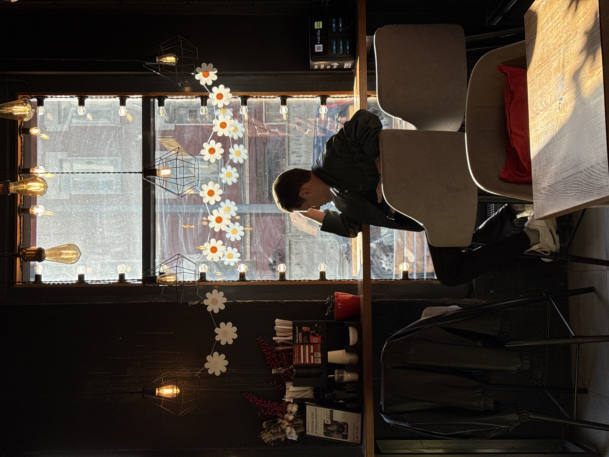 A person is seated at a table in a cafe. Outside the window is the city of Saint Petersburg, Russia. There are decorative daisies hanging in front of the window. The cafe has modern light fixtures. The table is next to a window.