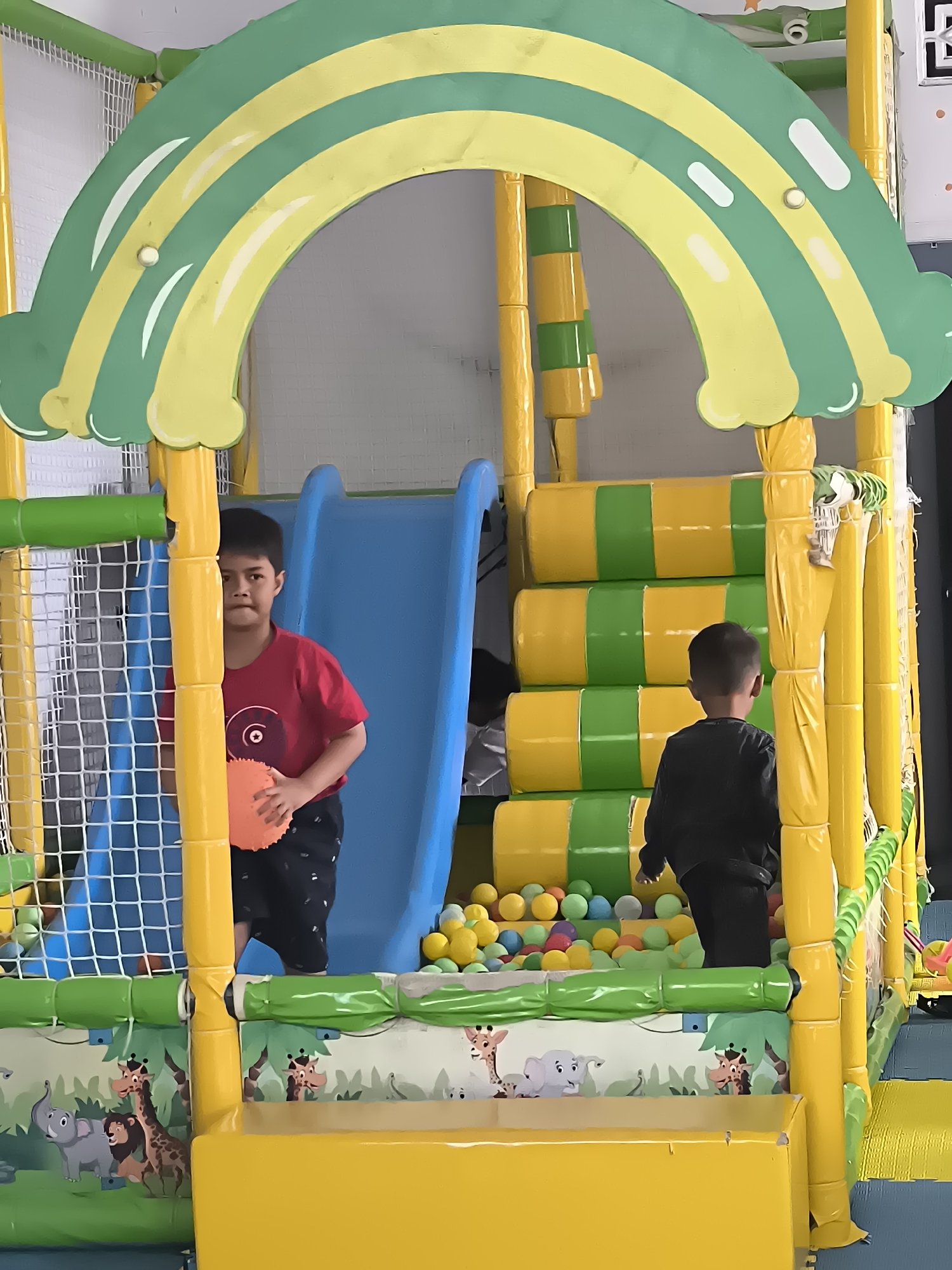 The image captures a vibrant indoor playground, likely located in Dagangan, Indonesia, featuring a large, colorful play structure designed with a rainbow motif at the top. The structure is primarily yellow and green, with a blue slide prominently displayed. Two young boys are the main subjects. The boy on the left, wearing a red t-shirt and black shorts, is positioned near the entrance to the play area, holding an orange circular toy. He is looking towards the camera with a curious expression. The boy on the right, dressed in a black long-sleeved shirt and pants, is further inside the structure, facing away from the camera, with his back to the viewer. The play area is filled with a large number of multi-colored plastic balls, visible in the ball pit area. The surrounding environment is enclosed, with net and padded walls forming the structure's boundaries. Decorative animal murals, including elephants, lions, and giraffes, adorn the lower parts of the play structure, suggesting it's a themed children's play area. The lighting suggests an indoor setting, possibly a dedicated children's entertainment venue.