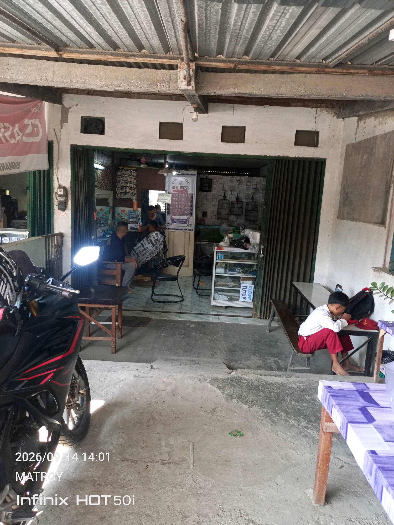 A street-level view shows the exterior of a barbershop in Semarang, Indonesia. A motorcycle is parked in the foreground on the left. Inside the shop, a barber is giving a haircut to a seated customer. Another person is standing behind the barber. To the right, a young boy in a white shirt and red pants is sitting on a bench, looking down at a table. The shop has a corrugated metal roof and an open front with folding doors. Shelves stocked with various items are visible inside. The ground is a concrete surface.