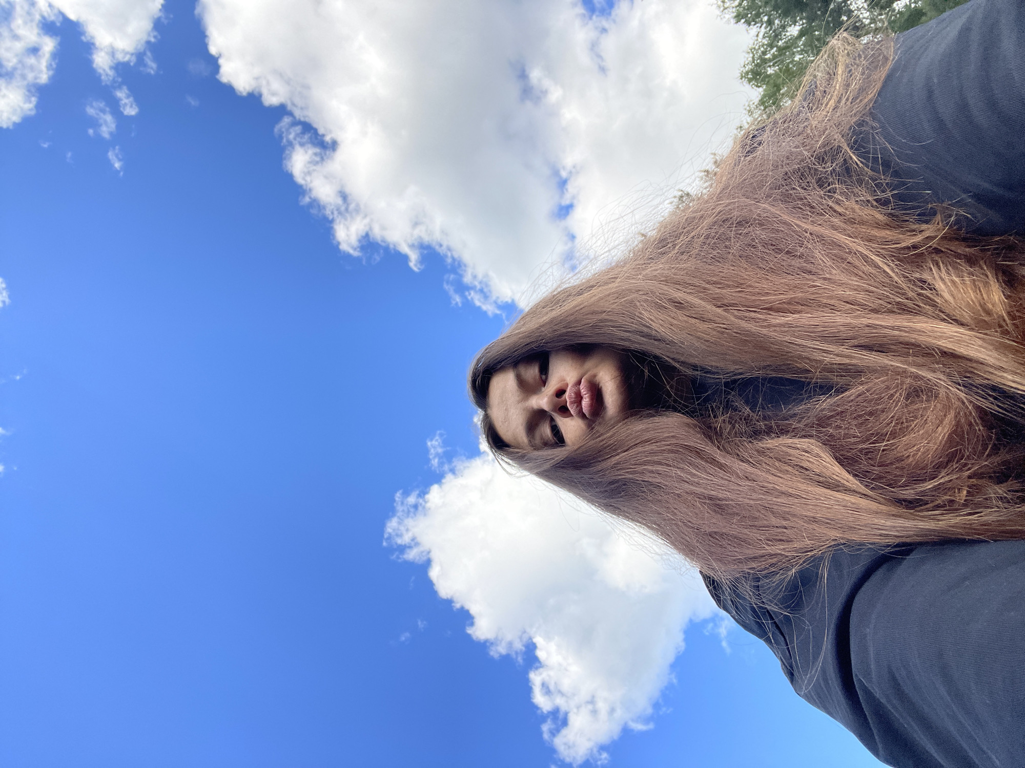 The image is a low-angle, selfie-style shot looking up at the sky. The primary subject is a young person with long, light brown hair that dominates the right side of the frame, obscuring much of their face. The person's lips are pursed, giving them a playful or slightly pouty expression. They are wearing a dark, possibly black, long-sleeved garment.
The background is a vibrant blue sky with scattered white, fluffy clouds. Some trees are visible in the upper right corner, suggesting an outdoor setting. The lighting indicates it is daytime, likely midday or afternoon, with bright sunshine illuminating the clouds and sky. The composition, with the camera pointing upwards, emphasizes the expanse of the sky and creates a sense of looking up at the person. The overall mood is casual and lighthearted. No text is visible in the image. The location context provided is Заречный, Russia.