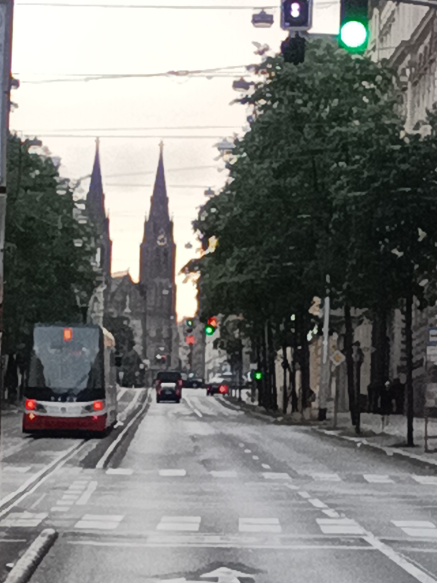 A tram, a red car, and several vehicles are on a street in Prague, Czechia. The tram is in the foreground on the left, moving away from the viewer. It is red and white with its headlights on. The red car is further down the street, also moving away from the viewer. The street has white lane markings and a pedestrian crossing. On the right side of the street are trees and buildings. In the background, two church spires are visible. Traffic lights are also present, with a green light illuminated. The sky is overcast.