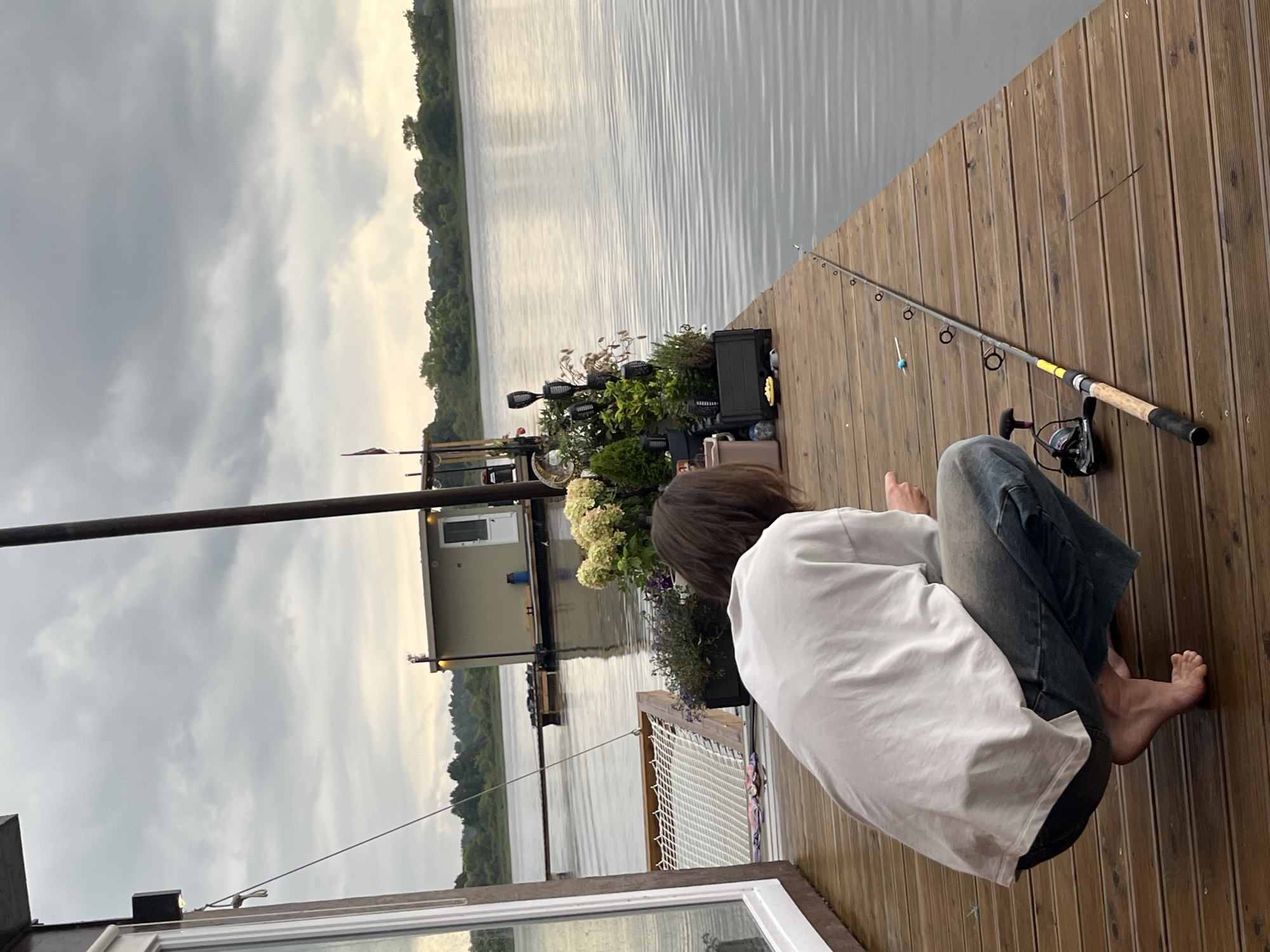 A person is crouching on a wooden dock, facing away from the camera, looking out at a body of water. They are wearing a white t-shirt and blue jeans. A fishing rod lies on the dock next to them, with its tip extending towards the water. In the background, a small houseboat is visible on the water, and a line of trees forms the distant shore. The sky is overcast with gray clouds. The scene appears to be set in Radchenko, Russia, on a calm, overcast day.