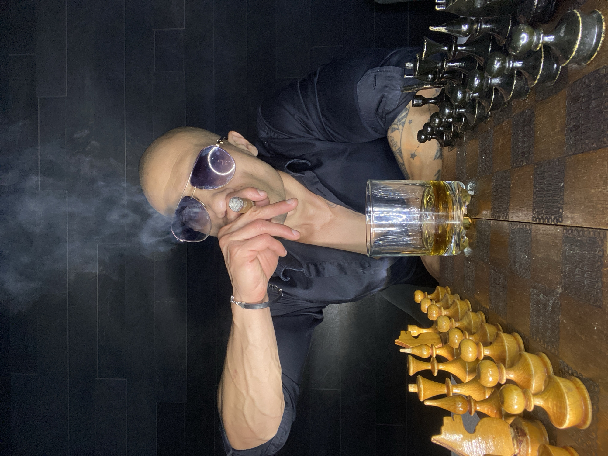 A man with a shaved head and sunglasses, dressed in a black shirt, is positioned next to a chessboard with chess pieces and a glass containing amber liquid. He holds a lit cigar, emitting smoke. The background is a dark, tiled wall. The location is not discernible from the image.