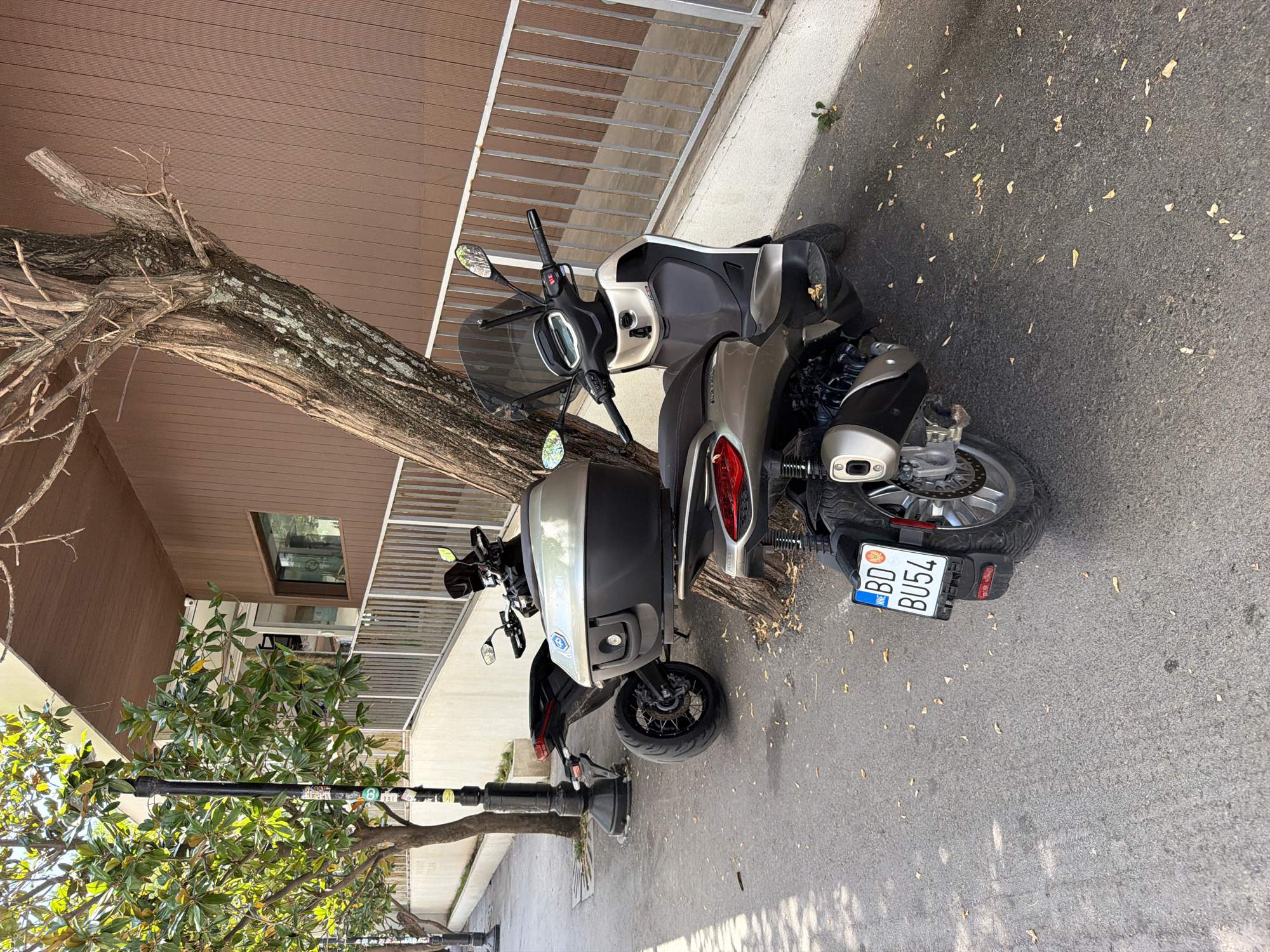 Two scooters are parked on a sidewalk next to a tree that leans against a building. The scooter in the foreground is silver and black, with a license plate that reads "BD BU54". It has a large storage box on the back. The scooter in the background is also silver and black, with a similar storage box. The building behind the scooters is covered in brown siding and has a metal fence in front of it. There are trees with green leaves on the left side of the image, and a lamppost with stickers on it. The ground is paved with asphalt and scattered with fallen leaves. The scene appears to be on a sunny day. The license plate "BD" is the city code for Budva, Montenegro.