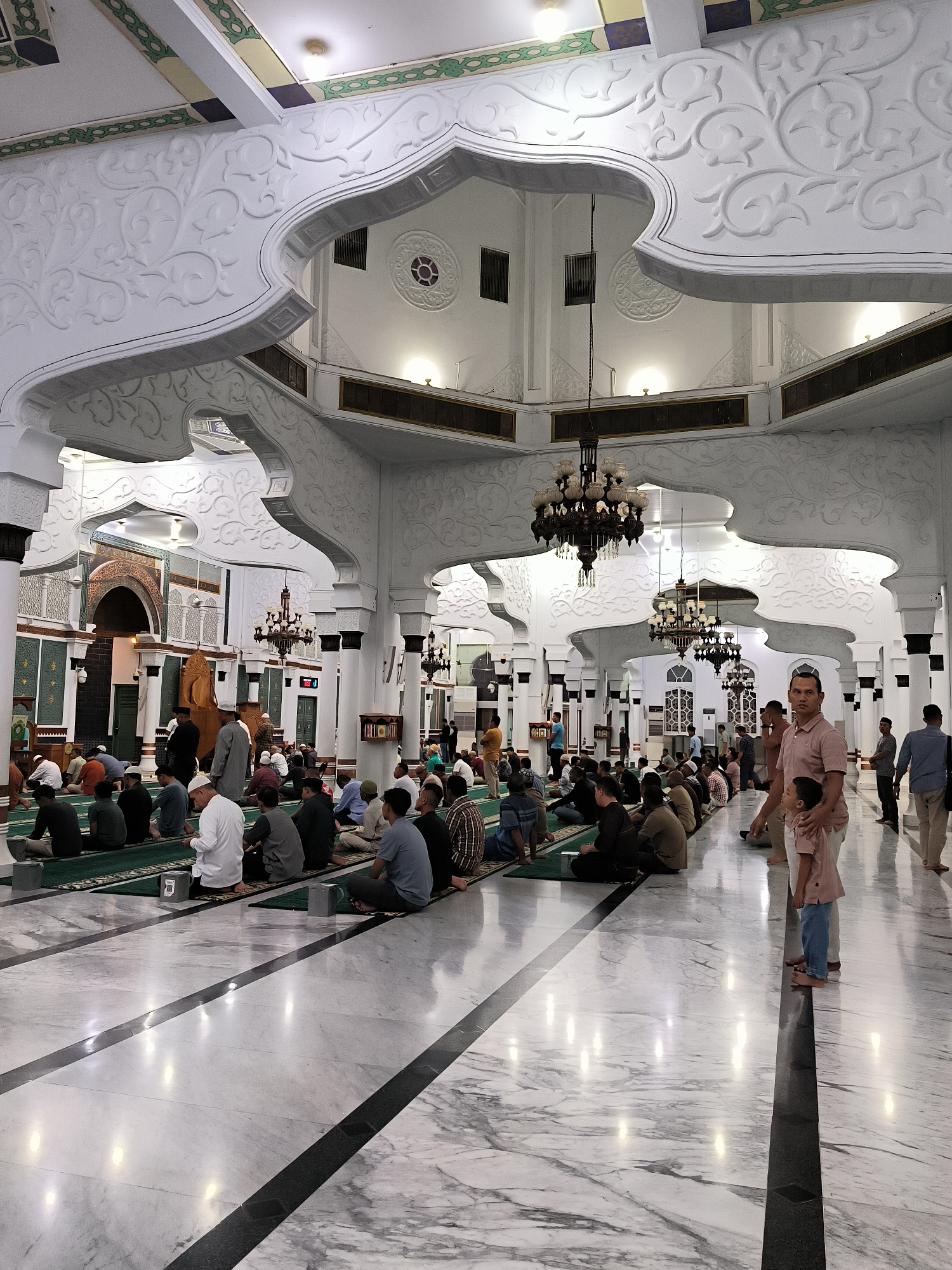 The image depicts the interior of a mosque, likely located in Banda Aceh, Indonesia, during prayer. A congregation of men is seated on green prayer mats arranged on the floor, facing towards the front of the mosque. The architecture is ornate, featuring white, intricately carved decorative elements on the walls and ceilings, and large chandeliers. The floor is a polished marble with a reflective surface. A man and a young child are standing in the foreground, looking towards the congregation. Several other individuals are visible in the background, some standing and others seated. The scene suggests a communal prayer or gathering within a significant religious space.