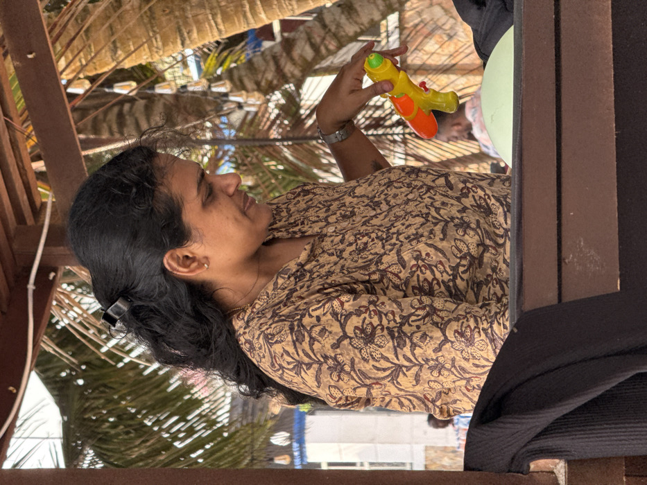 An image taken in Mopo Onijebu, Nigeria, shows a woman in profile, positioned under a wooden overhead structure. Her dark, wavy hair is pulled back with a black claw clip. She wears a light brown, long-sleeved shirt patterned with dark floral and leaf motifs. A thin gold chain is visible around her neck, and a silver-toned watch or bracelet is on her left wrist. Her right hand holds a yellow and orange toy water gun with a green nozzle, pointed downwards. In the background, out-of-focus palm trees and string lights with clear bulbs are visible. Indistinct figures and part of a light-colored building can be seen further in the distance. The foreground features a dark, ribbed fabric on the lower left and a light green, possibly inflated, object on the lower right, both resting on a dark wooden surface.