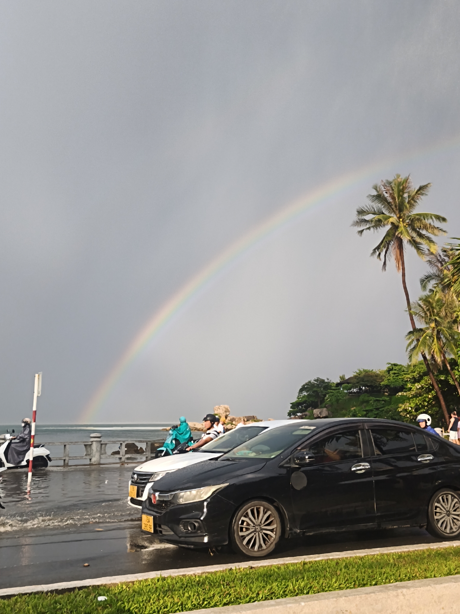 The image depicts a street scene with a prominent rainbow arching across a cloudy sky. In the foreground, a black sedan is parked on a wet road, with its front end angled towards the viewer. To its left, a white sedan is also visible, partially obscured by the black car. Further down the road, a person on a turquoise scooter, wearing a blue rain poncho, is riding away from the camera. A white scooter with a rider covered in a black poncho is also present on the left side of the image. The road appears to be flooded in some areas, with water splashing around the wheels of the scooters. In the background, a low stone wall separates the road from the sea, and beyond that, a lush green landscape with palm trees and vegetation can be seen on a hill. The overall atmosphere suggests a recent or ongoing rain shower.