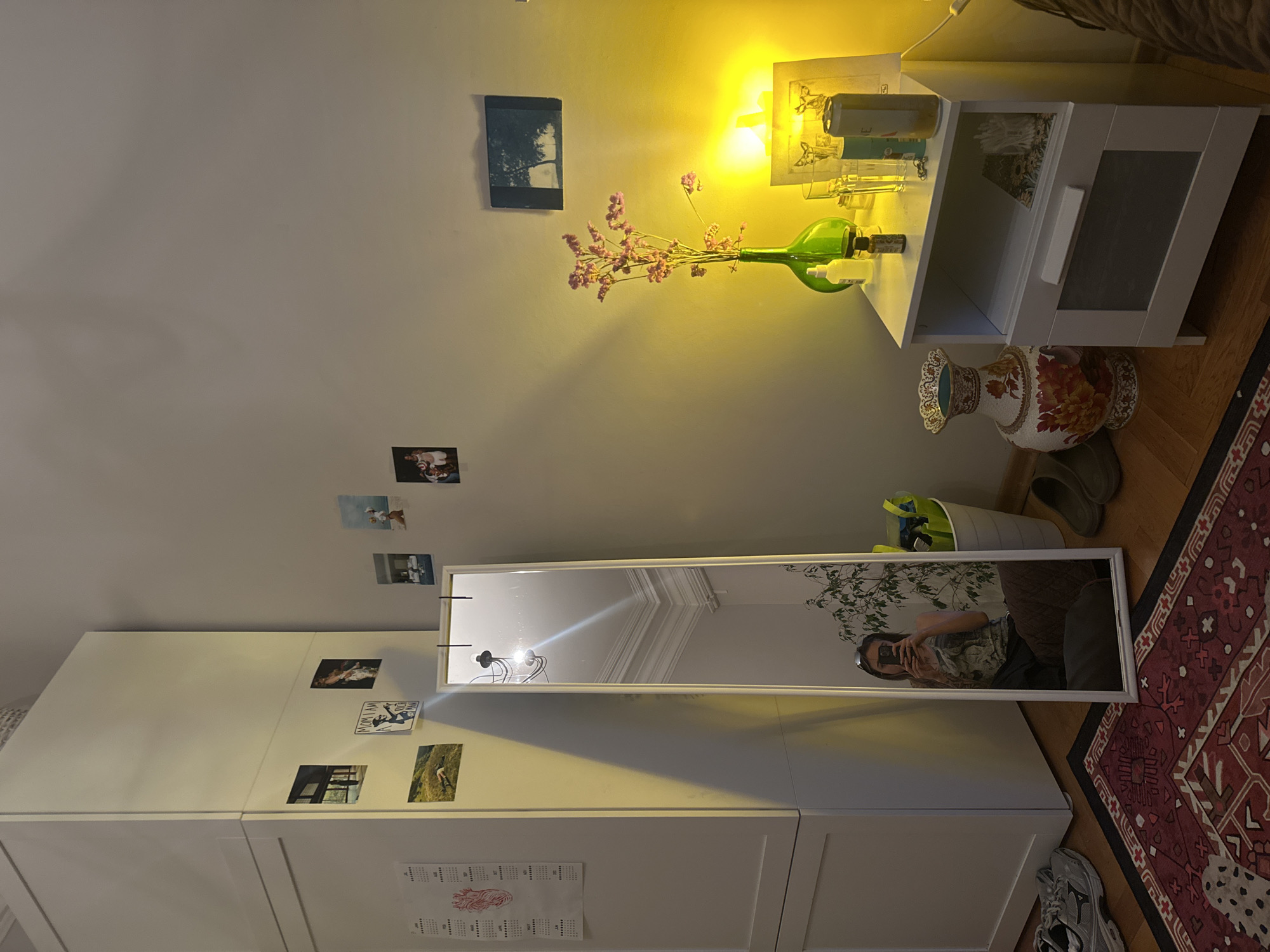 The image shows a bedroom interior, likely in Moscow, Russia, bathed in warm, artificial light. A tall, white wardrobe stands against the left wall, adorned with several small framed photographs and calendars. A full-length mirror leans against the wardrobe, reflecting a woman taking a selfie, a potted plant, and the ceiling. To the right of the mirror, a white nightstand holds a variety of items: a green vase with dried flowers, a framed picture, several glasses, a bottle, and a can. A large, ornate vase sits on the floor next to the nightstand. The floor is covered with a patterned rug and wooden planks. A pair of grey sneakers rests on the rug to the left, and a pair of dark shoes is positioned to the right.