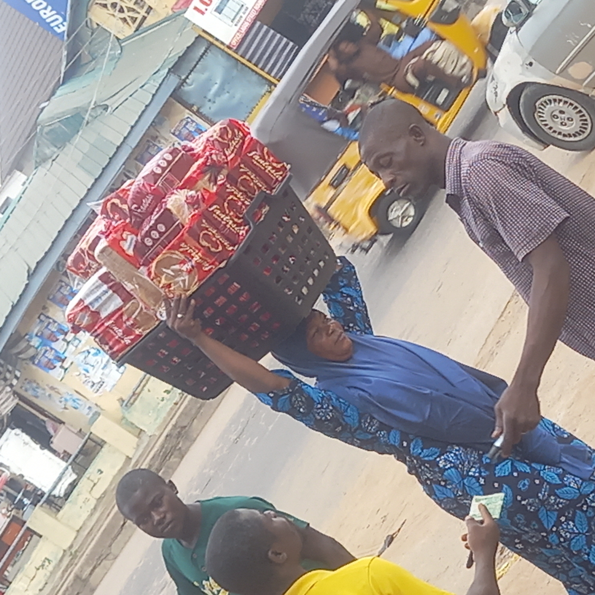 This image captures a vibrant street scene, likely a market or commercial area, in Sardauna, Nigeria, during daylight hours. The central focus is a woman who skillfully balances a large, black plastic basket filled with packaged bread on her head. She wears a blue head covering (hijab) and a patterned blue dress, with her arms raised to steady the heavy load.
She appears to be a street vendor, interacting with customers. A man in a red and white checkered shirt leans towards her, possibly engaging in a transaction or conversation. In the foreground, two younger individuals are visible. One, wearing a green shirt, looks towards the camera, while another in a yellow shirt is seen with his hand extended, holding what appears to be money, suggesting a purchase from the woman. A small currency note is visible in the woman's hand.
The bread packages, predominantly red, clearly display the brand name "Fantastica" and possibly "Fast food." In the background, typical urban elements include a yellow auto-rickshaw (keke Napep) with occupants and a silver car, indicating a busy thoroughfare. Buildings line the street, with partial views of signs. One sign has "EURO" written on it, and another shows "I.G." followed by "OFFICE HOURS." The overall scene depicts a common daily life interaction in a bustling urban environment.