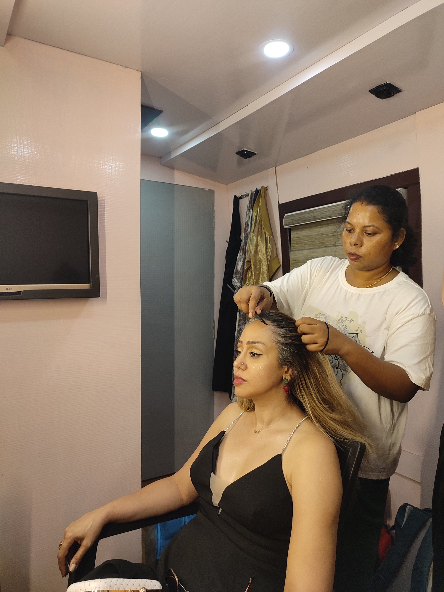 In an indoor setting, likely a dressing room or salon in Kalamassery, India, a dark-skinned woman with her hair pulled back and wearing a white graphic t-shirt and a gold chain is styling the hair of another woman. The stylist, who has a bindi on her forehead, is carefully working on the roots and sections of the seated woman's long hair with her hands, possibly using a hair clip or pin.
The woman being styled has light skin, long, light blonde or light brown hair, with noticeable grey roots. She is wearing a black V-neck top with thin, sparkly shoulder straps. Her makeup includes prominent winged eyeliner, and she has a nose ring and red and green dangle earrings. Her left arm, adorned with a ring, rests on the black armrest of her chair. Both women appear focused and calm.
The room features light beige, textured walls and a white ceiling with multiple recessed lights. A black LG flat-screen television is mounted on the left wall. In the background, behind the standing stylist, clothes hang on a rack, including a black garment and a gold textured fabric. There's also a frosted or tinted glass panel or door, and a window or opening with a dark wooden frame and a beige blind. Two small, black, ceiling-mounted devices, possibly cameras or sensors, are visible. The overall atmosphere suggests preparations for an event or photoshoot.