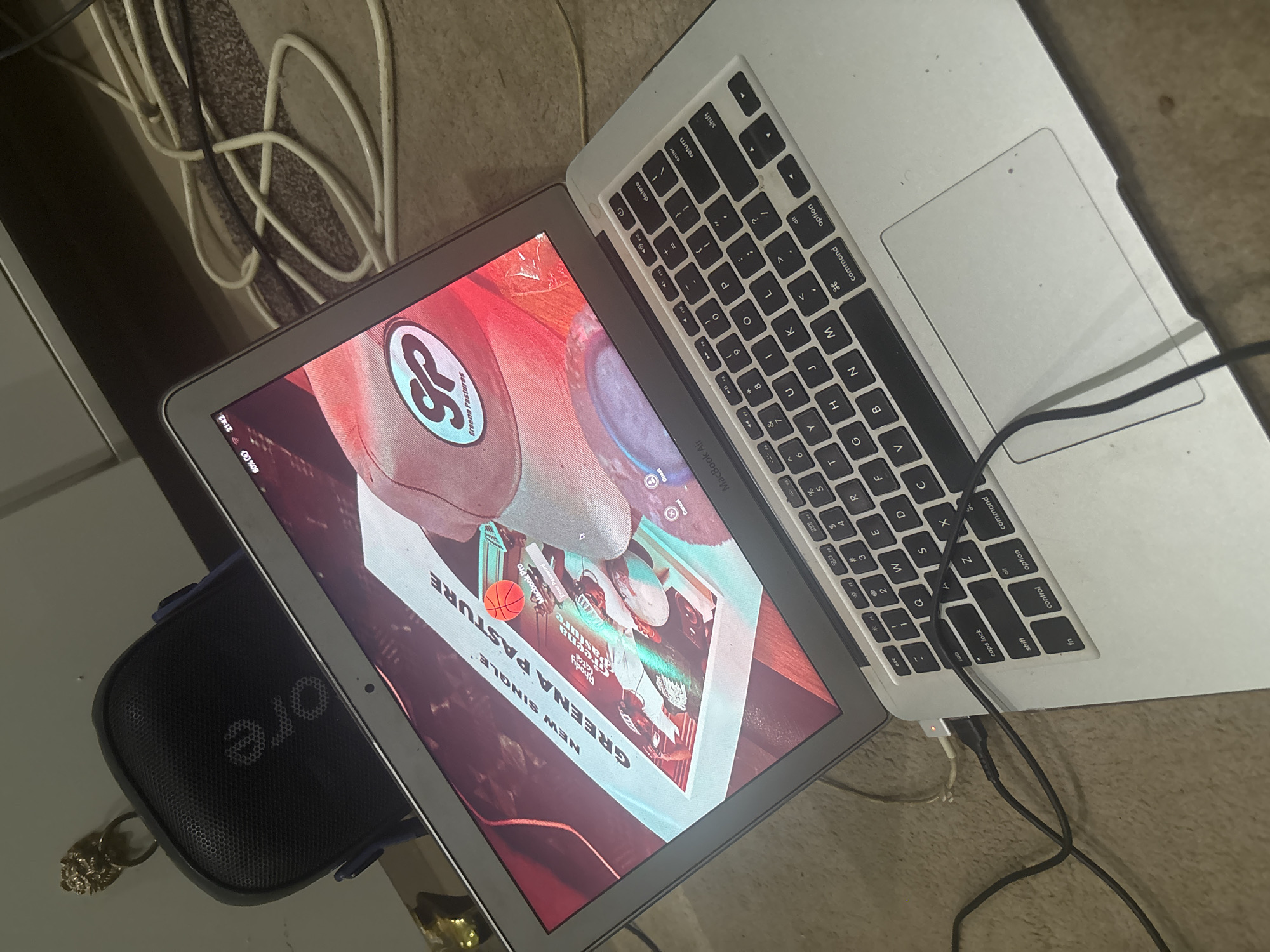 The image shows a laptop displaying album art for "Greena Pasture" by SP, with a basketball. The laptop is on a beige floor. To the left of the laptop is a black Bluetooth speaker with an attached keychain with a lion's head. A white cable is present, along with black charging cables connected to the laptop. The image provides no information about Abuja, Nigeria.
