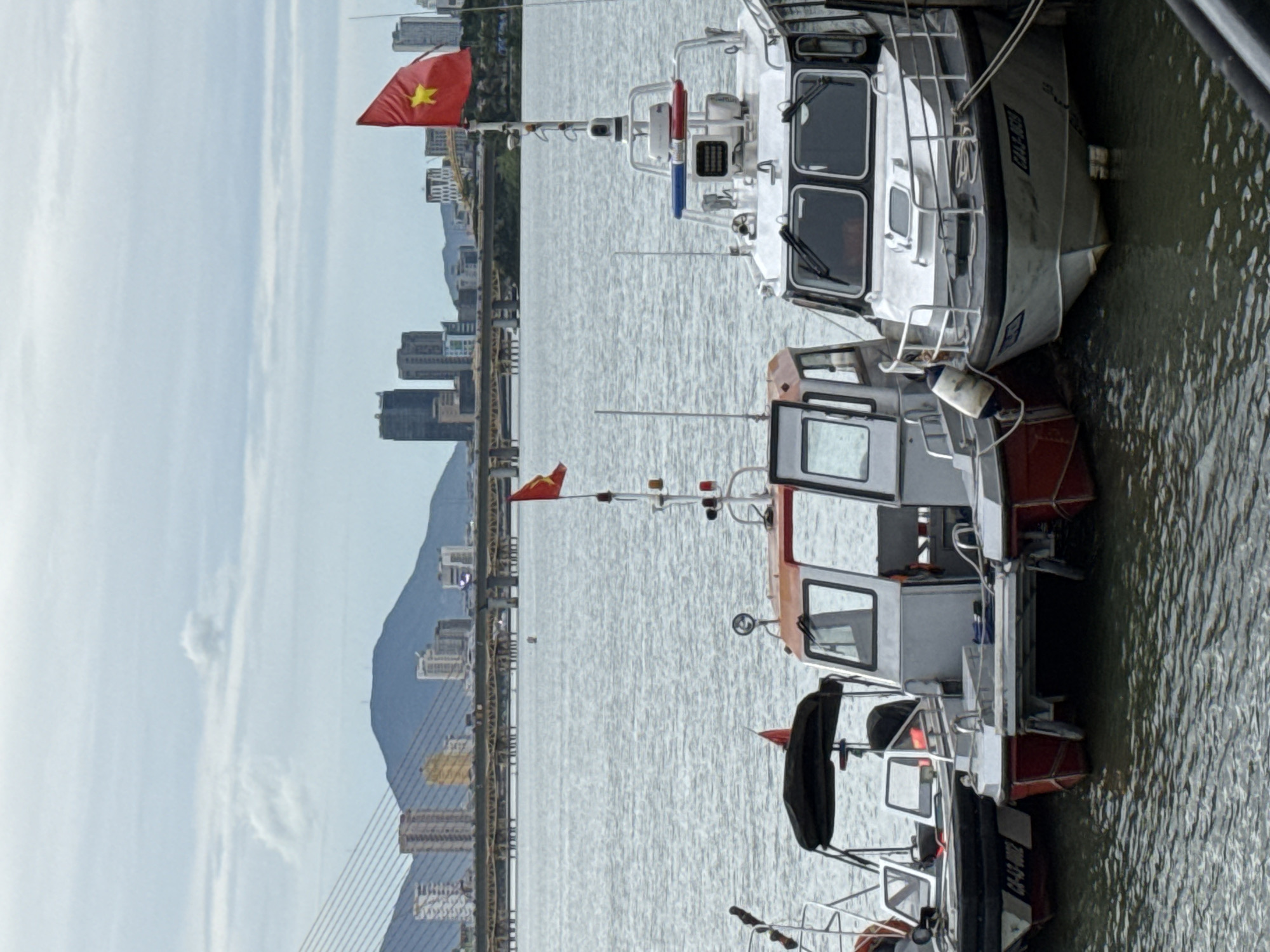 A slightly overcast day with calm waters shows several boats docked near a city skyline. In the foreground, three boats are tied together. The leftmost boat is white with a black canopy. The middle boat is white with an orange cabin and a small red flag. The rightmost boat is a white patrol boat with a blue and red light bar on top and a Vietnamese flag flying from its mast. The boat has the registration number "DN1309" on its hull. In the background, a modern city skyline with tall buildings and a bridge under construction is visible, with mountains beyond. The scene appears to be a waterfront area in Đà Nẵng, Vietnam, likely near the Han River. There are no people visible in the image.