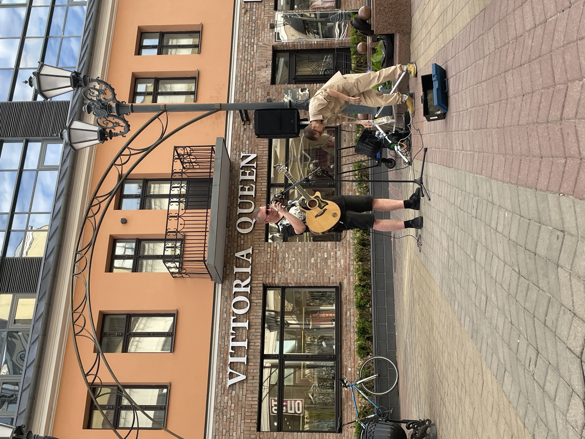 In the city of Brest, Belarus, on a paved street, a street musician is performing with an acoustic guitar. He is wearing a black t-shirt, black shorts, and black socks with sneakers. He is also wearing sunglasses. To his right, another person in a beige jumpsuit is attending to some equipment, bending over and holding a bottle of water. The background features a building with an orange facade and a brick lower level. The building has large windows and a balcony with an ornate railing. Above the windows, the text "VITTORIA QUEEN" is prominently displayed in large silver letters. A bicycle is parked to the left of the musician, and a street lamp is visible in the upper right of the frame. The scene appears to be an outdoor commercial area, likely a pedestrian zone, given the paved walkway and the presence of storefronts.