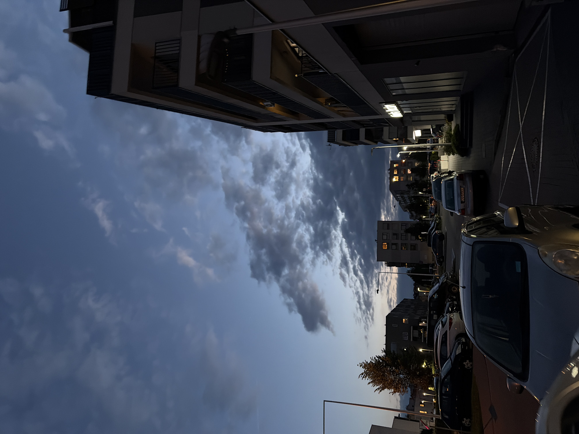 This image depicts a twilight scene in a residential area of Lublin, Poland. The sky is a deep blue with scattered, textured clouds, suggesting the end of daylight. In the foreground, the front of a silver car is visible, its surface reflecting the dim light. Behind it, a parking lot is filled with various cars, including a black hatchback, a white SUV, and a dark-colored van.
To the right, a modern apartment building with multiple balconies and dark railings dominates the frame. The building's facade is light-colored, and some windows are illuminated, indicating occupancy. A street lamp stands near the building.
In the background, more apartment buildings are visible. One building on the left has a few illuminated windows, and a tall, slender street lamp stands in front of it. A single tree with autumn foliage is situated between buildings on the left. Another apartment building further in the background has several glowing windows. The overall impression is that of a quiet urban evening in Lublin.