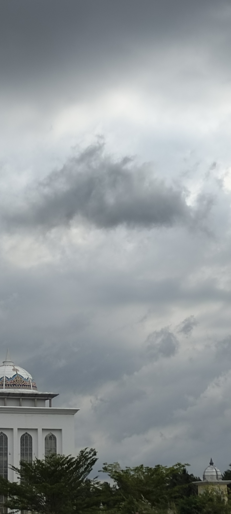 The image displays an outdoor scene under a heavily overcast sky, likely in Hueng, Indonesia. The sky is dominated by dense, dark gray clouds, suggesting a gloomy day or impending rain.
In the lower left and center, a portion of a prominent white building is visible, characterized by a large, ornate dome. This dome features intricate patterns in blue, white, and muted orange tones. Below the dome, arched windows are visible along the building's facade, indicative of Islamic architectural style. Green trees with dense foliage are prominent in the foreground and midground, partially obscuring the lower sections of the building. Further to the right, nestled amongst the trees, a smaller, simpler dome of another structure can be seen.
No people or activities are present in the frame, presenting a static and serene scene. The time of day appears to be daytime, though the heavy cloud cover creates subdued lighting. There is no visible text. The architecture strongly suggests the primary building is a mosque or a similar religious institution.