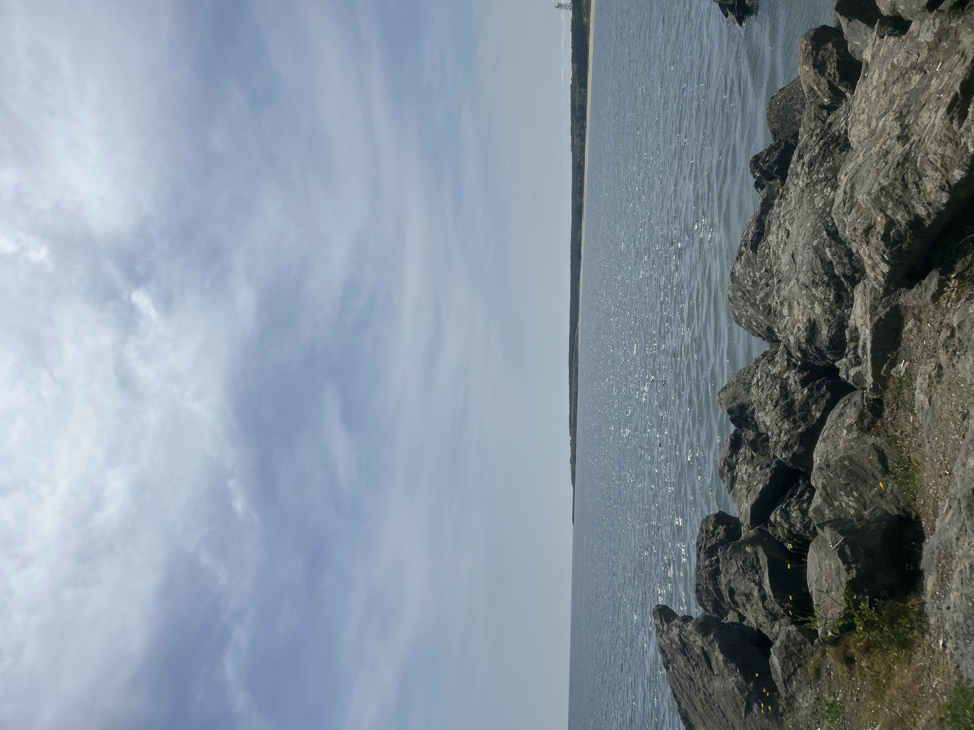 The image depicts a coastal scene with the sky taking up approximately half of the view on the left side. The sky displays a mix of light blue with wispy white clouds. The right side of the image is dominated by a body of water, its surface shimmering with reflected sunlight. Along the water's edge are large, gray rocks. In the distance, across the water, is a thin strip of land, possibly a beach or coastline, with a few structures visible near the far end. The location is likely Rezovo, Bulgaria. There are no visible people or apparent actions. The environment consists of the sky, water, rocky shoreline, and distant coastline.