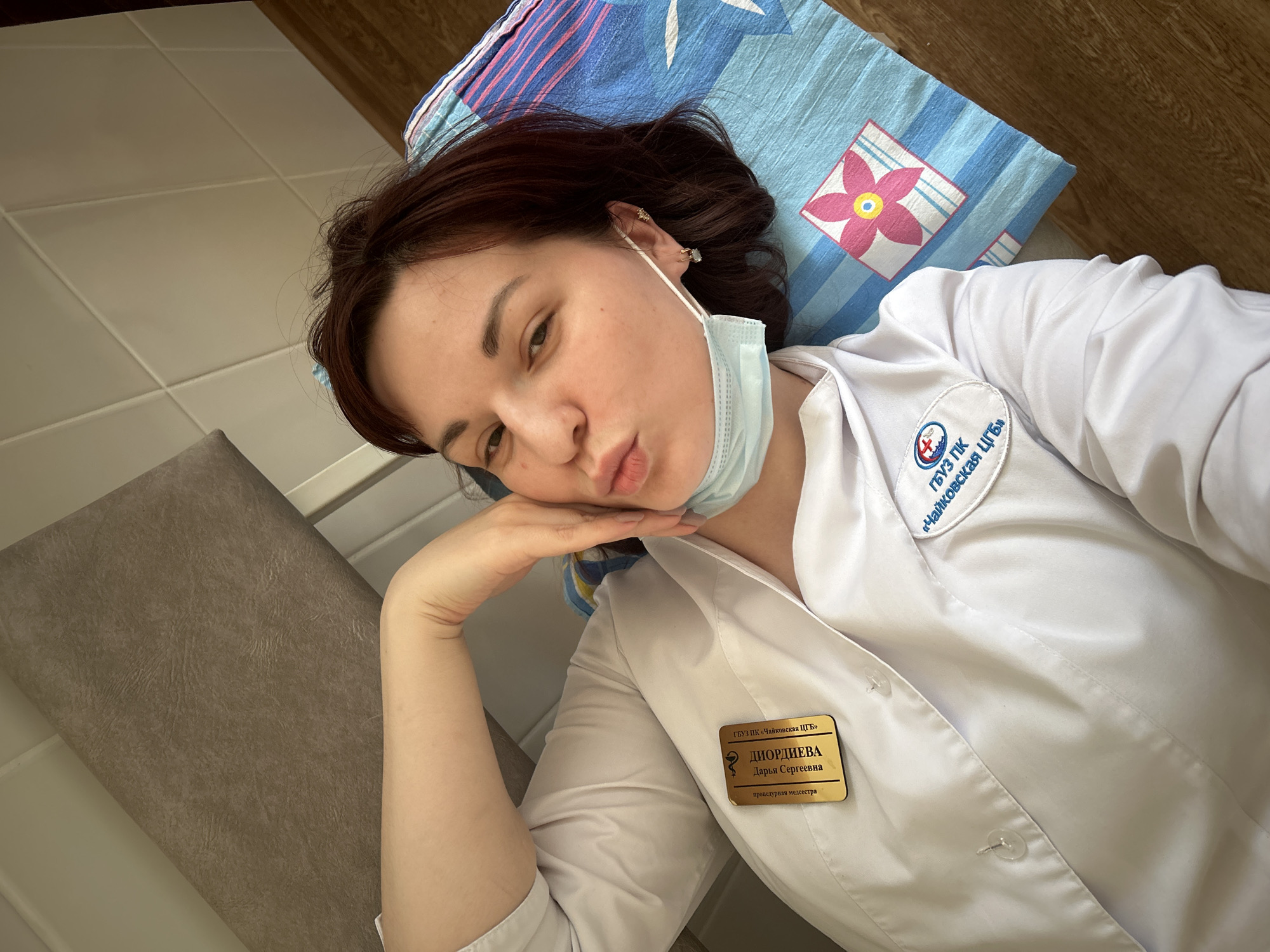 A medical professional, identified as Darya Sergeevna Diordieva, a procedural nurse, is captured in a selfie. She is lying down, propped on her left arm, and making a playful, slightly pouty expression towards the camera. Her dark hair is spread around her head, which rests on a blue and white patterned pillow featuring a pink and yellow flower design. A blue surgical face mask is pulled down, resting under her chin.
She is wearing a white medical lab coat. On her left chest, a circular emblem is visible, bearing the text "ГБУЗ ПК «Чайковская ЦГБ»" (State Budgetary Healthcare Institution of Perm Krai "Chaikovskaya Central District Hospital") along with a red cross symbol. Below this, a gold name tag provides her full name, "ДИОРДИЕВА Дарья Сергеевна," and her profession, "процедурная медсестра." A patterned blue and yellow scarf is also visible near her neck.
The setting appears to be an indoor medical facility. To her left, there's a brown, textured surface that looks like a padded medical bench or chair. The background shows a light-colored tiled wall and a wooden floor. The image conveys a casual, perhaps tired or lighthearted, moment from a medical professional, likely taken during a break in a facility within or near Фоки, Russia, given the "Chaikovskaya Central District Hospital" identifier.