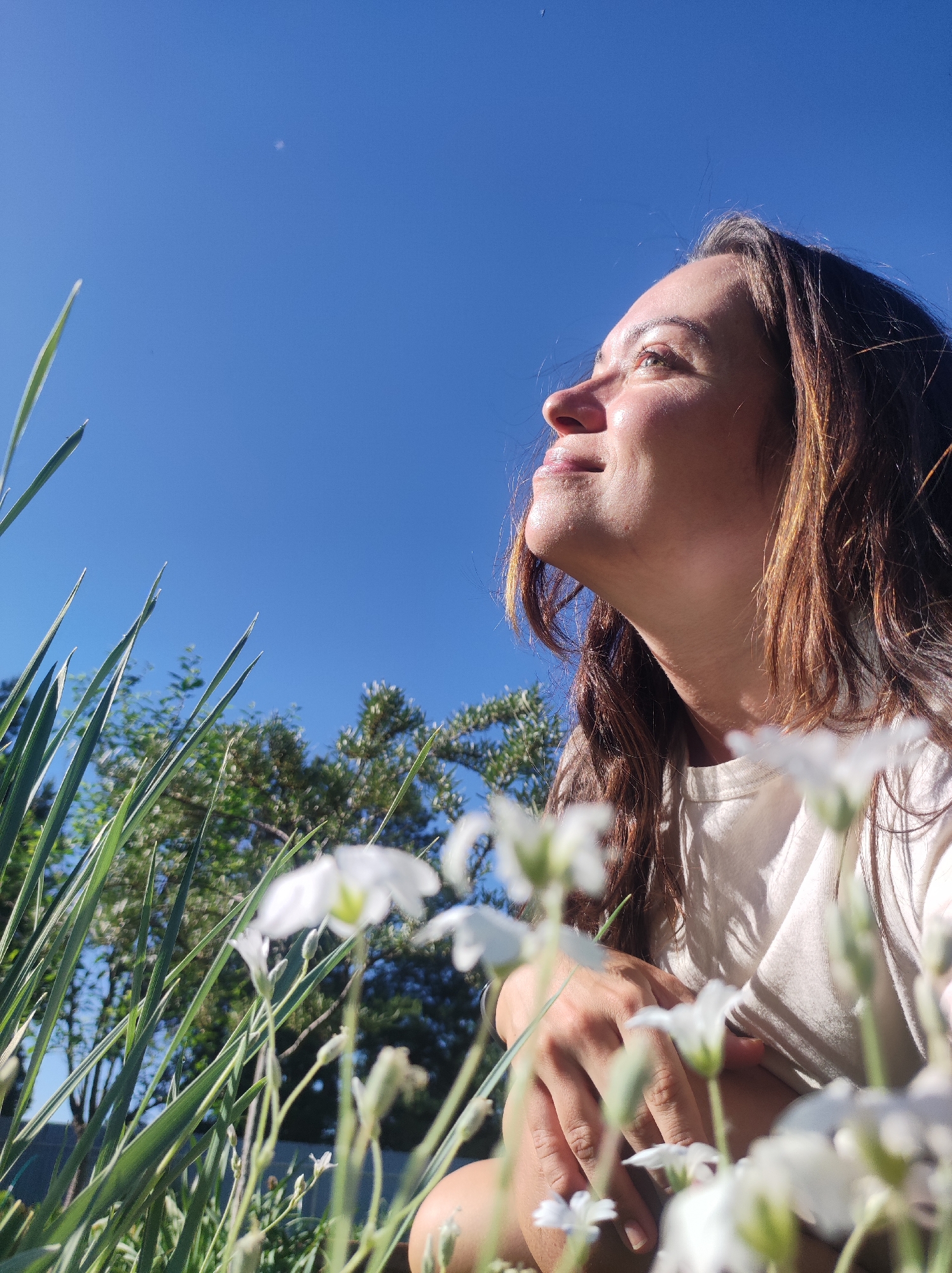 The image depicts a woman with long, brown hair looking up at a clear blue sky. She is positioned in the foreground, with the camera angle looking up from a low perspective. In front of her are out-of-focus white flowers and green grass, suggesting she is outdoors in a garden or natural setting. The woman is wearing a light-colored t-shirt. The background shows trees and a fence, hinting at a residential or semi-rural environment. There is no information in the image to indicate a specific location within the city of Фоки, Russia.