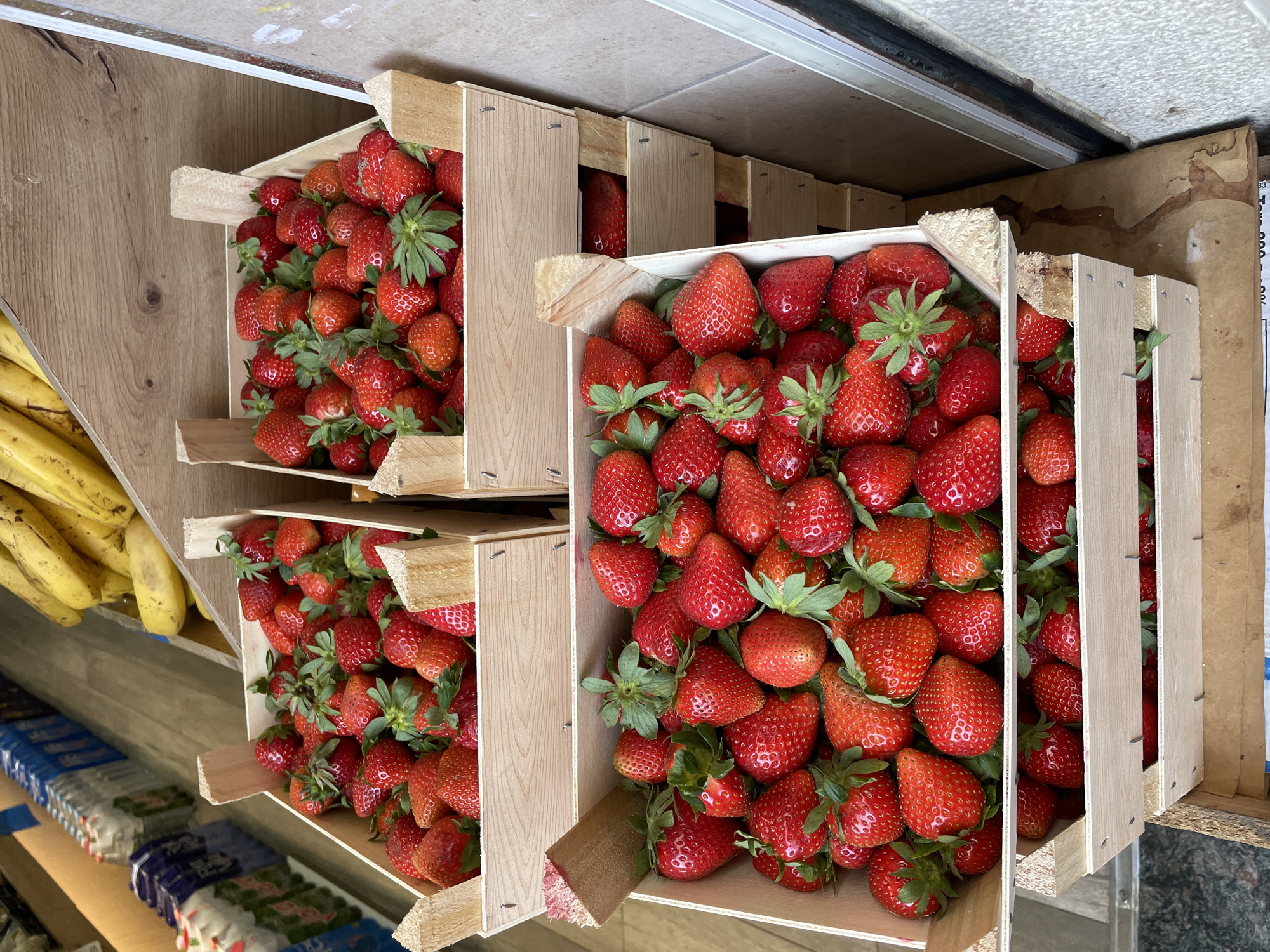 The image shows three wooden crates filled with strawberries. The crates are stacked on a shelf, with a bunch of bananas visible to the left of the top crate. Below the bottom crate, there are rows of what appear to be packaged goods on a shelf. The image appears to be taken in a market or shop. The location is likely in the city of Barcelona, Spain, based on the image context.