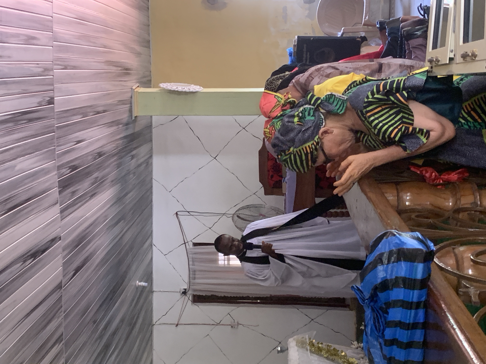 An indoor scene in Ukwu-Abwa, Nigeria, shows an elderly woman in African print clothing and headwear leaning forward with hands extended over an ornate wooden structure covered with clothing and a blue striped bag. A man in a white robe with black trim stands behind the woman. The room has white walls with cracked patterns, a fan on the wall, and a tiled wall to the side.