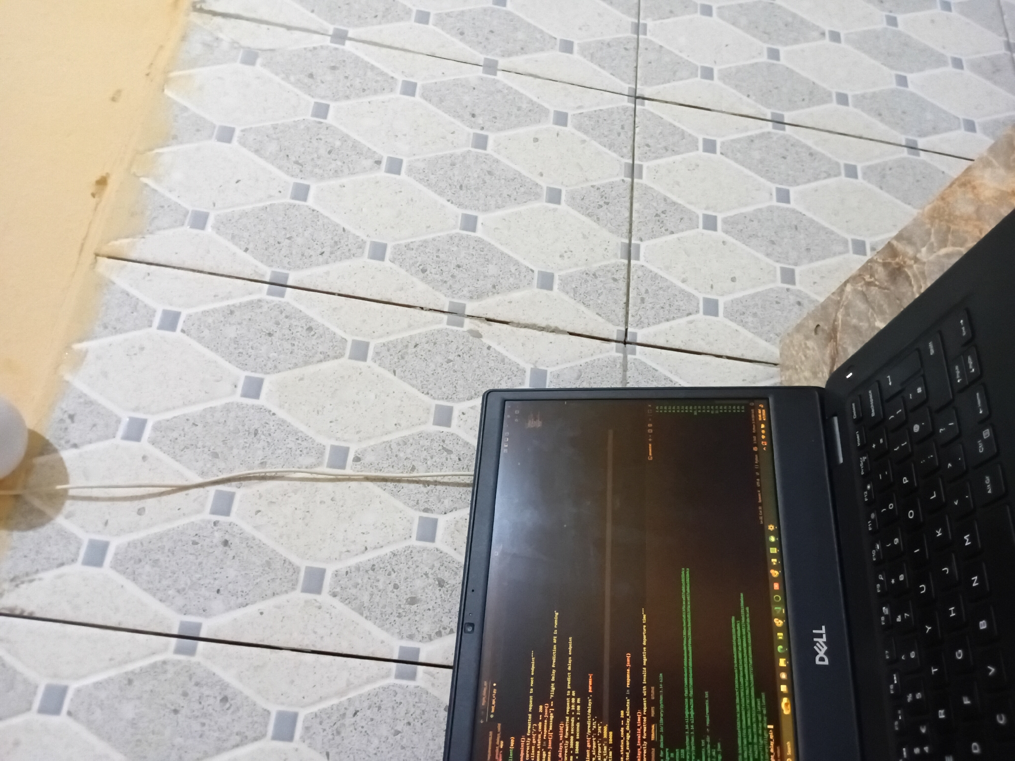 The scene depicts a Dell laptop resting on a tiled floor. The laptop screen displays code related to a "Flight Delay Prediction API." The code snippet includes functions for handling requests, validating delays, and managing airport and time parameters. The laptop keyboard is visible to the right. The floor consists of gray and white hexagonal tiles with a pattern. A white electrical cord is partially visible in the background. The overall setting appears to be indoors.