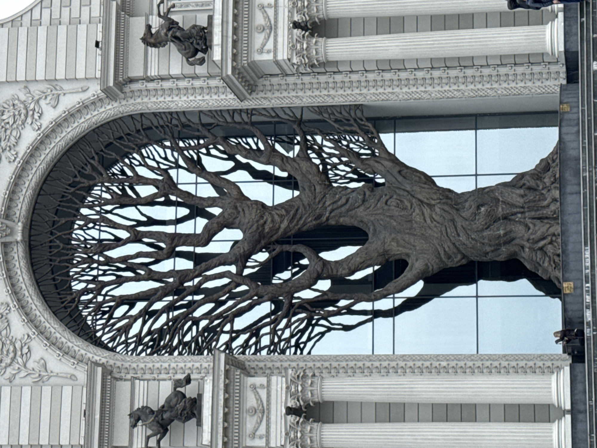 A detailed architectural close-up reveals a grand building facade dominated by a massive, sculptural representation of a gnarled tree or intertwined roots, framed within a large, ornate oval window. This dark, intricately branched sculpture forms a striking silhouette against the light, possibly sky-reflecting glass panes behind it, giving the impression of a colossal tree integrated directly into the building's design.
The facade is richly adorned with classical and highly decorative elements, including light-colored fluted columns, elaborate cornices with repeating patterns, and decorative friezes featuring leaf motifs and other ornamentation. Several smaller, dark-colored equestrian statues are integrated into the facade; one is partially visible in the upper left corner, depicting a rider on a rearing horse, and another is more clearly seen in a lower right niche, showcasing a similar dynamic pose.
In the upper right corner, the lower body and legs of a person are partially visible, suggesting an activity like maintenance or construction on the building's exterior. The scene is illuminated by bright daylight, highlighting the detailed artistry of the structure. The overall impression is one of elaborate grandeur and a blend of natural and architectural forms. No discernible text is visible in the image. The setting is a public building in Kazan, Russia, known as the Palace of Farmers.