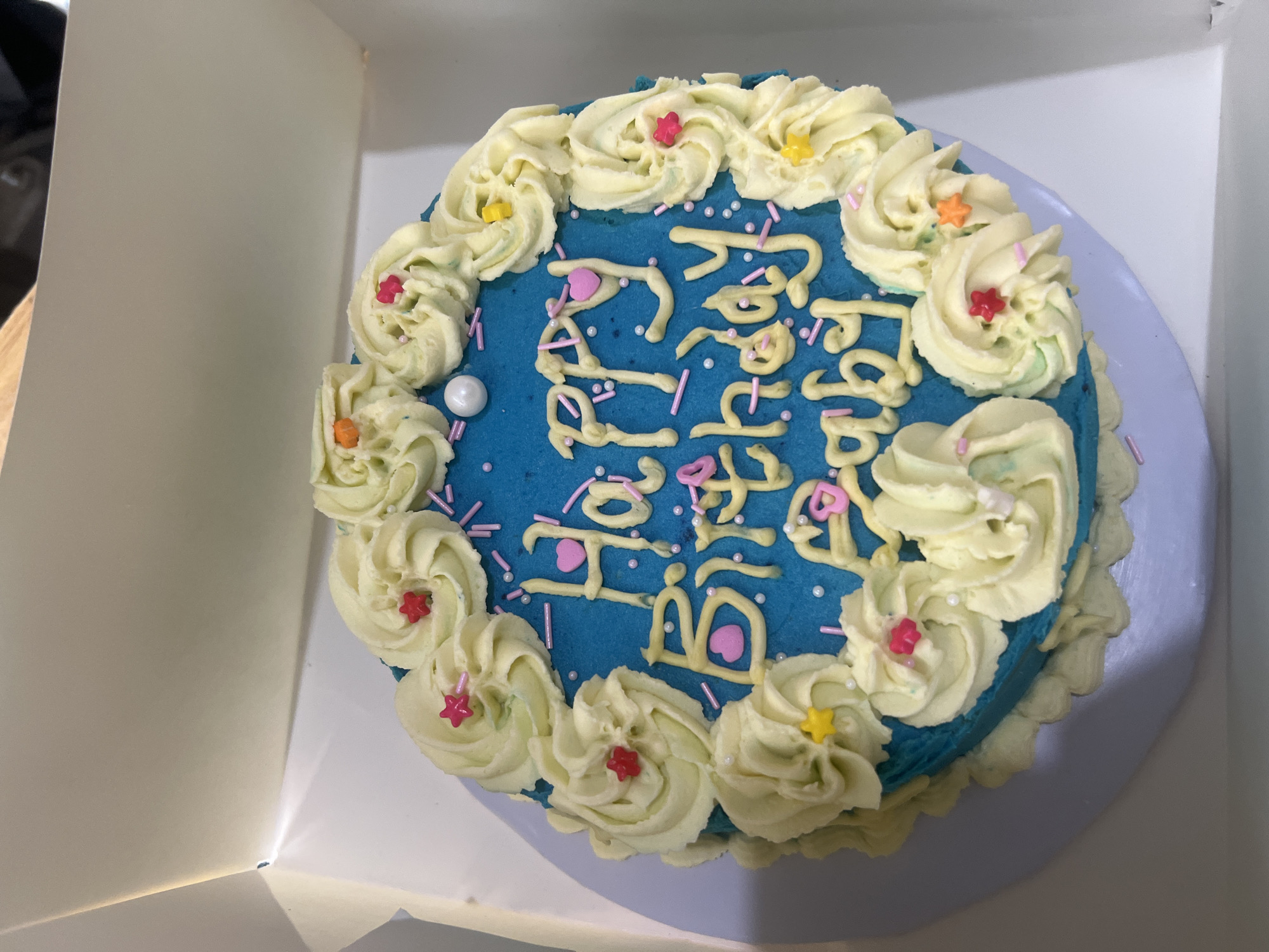 The image shows a blue-frosted birthday cake with off-white frosting rosettes around the edge. The cake is decorated with pink sprinkles and a yellow frosting inscription, which reads "Happy Birthday". There are pink heart-shaped decorations on the cake, and small star-shaped sprinkles on top of the rosettes. The cake sits on a white plate inside a white cardboard box. The scene appears to be indoors. There is no information in the image that would suggest this image was taken in Jos, Nigeria.