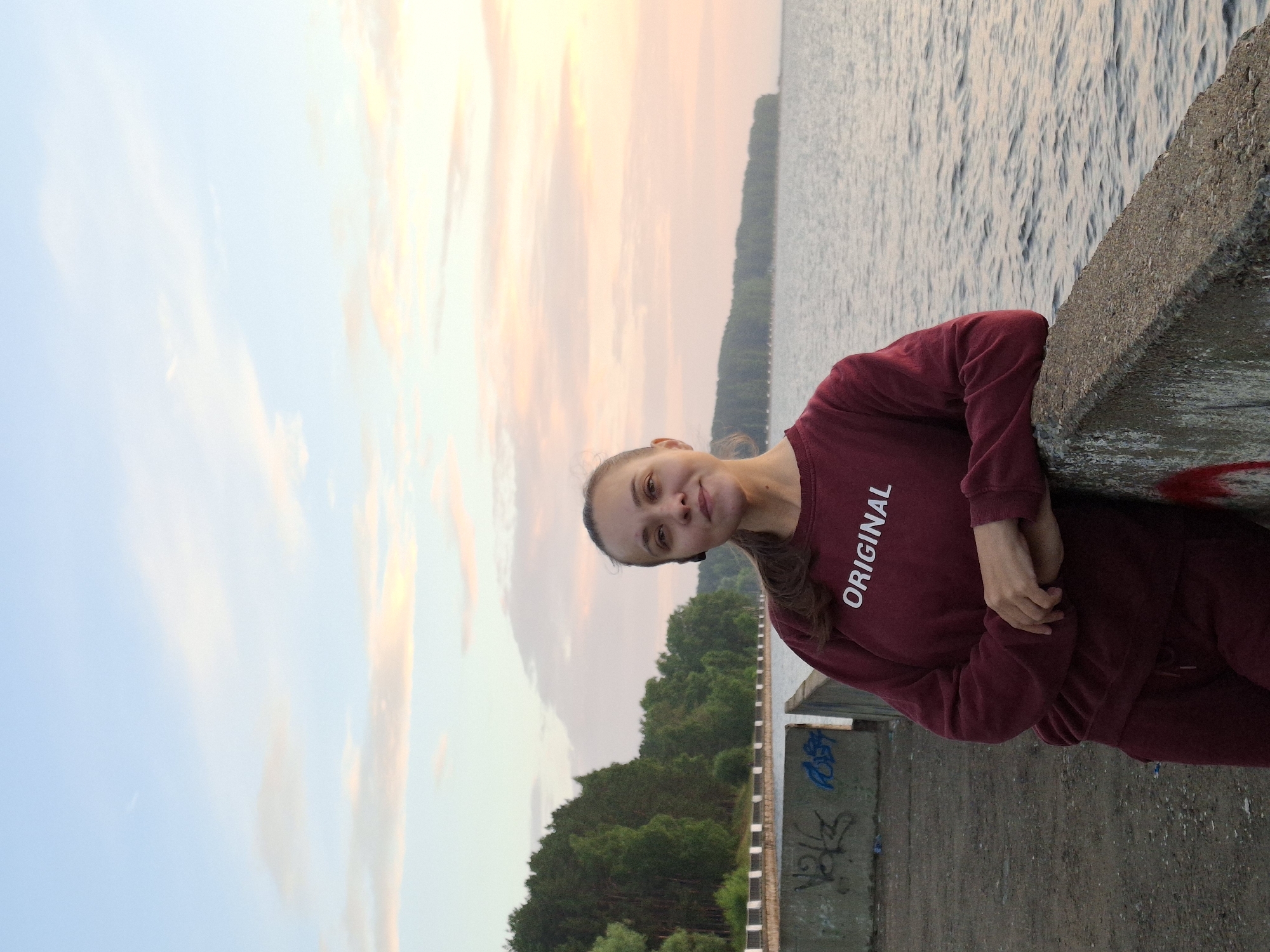 A young woman with brown hair pulled back in a ponytail, wearing a maroon sweatshirt and matching pants, stands by a concrete barrier overlooking a body of water. The sweatshirt has "ORIGINAL" printed in white text across the chest. She is wearing a black Bluetooth earpiece. The background features a tree-lined shore and a pale sky with streaks of pink and orange clouds, suggesting a sunset. The concrete barrier has graffiti on it. The location is on the embankment of the Kama River in the city of Chaikovsky, Russia.