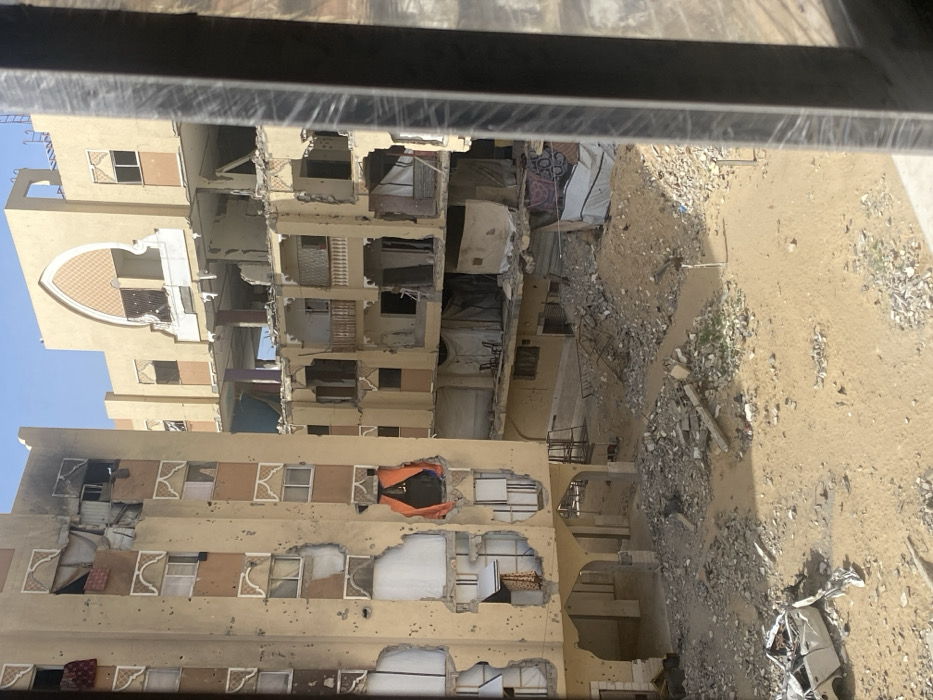 The image depicts a devastated urban landscape in Khan Yunis, Palestinian Territory, under a clear blue sky. The scene is dominated by multiple multi-story residential buildings exhibiting extensive damage.
In the foreground, a sandy, unpaved ground is covered with piles of concrete rubble, rebar, and shattered building materials. A heavily damaged white car, possibly a sedan, is visible on its side in the lower-left, crushed and largely disassembled.
To the left, a multi-story building, light brown in color, shows numerous pockmarks consistent with bullet or shrapnel impacts across its facade. Several large sections of its exterior walls are blown out, exposing interior rooms with dangling wires, fabric, and debris. Windows are shattered or missing, with some openings covered by makeshift tarps or fabrics, including a prominent orange sheet in a lower-mid-level opening.
In the center and extending to the right, another set of interconnected buildings, also light brown, features significant structural collapse. Large portions of upper floors and balconies are missing or collapsed inward, leaving internal structures exposed. Intact elements reveal decorative arched window frames and a large ornate arch on the uppermost visible section. In one exposed lower-level room, two light-colored chairs are visible amidst the debris.
The perspective is from an elevated position, looking out through what appears to be a window frame, with a dark, vertical structural element partially obstructing the view on the far right. No people are visible in the scene.