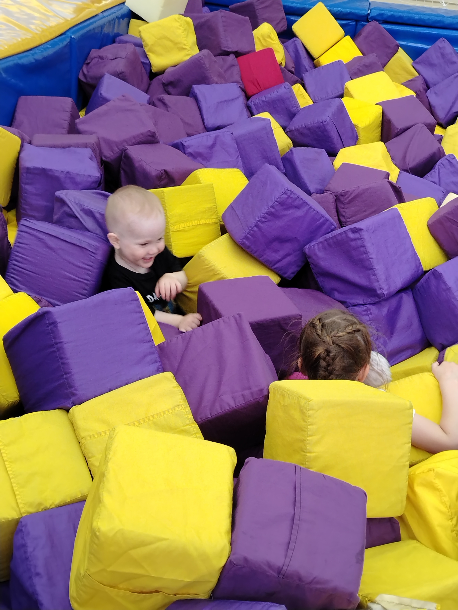 This image depicts an indoor recreational foam block pit, located in the city of Chaikovsky, Russia. The pit is a large, rectangular enclosure with its upper perimeter lined with blue protective padding. It is densely filled with numerous soft, fabric-covered blocks, primarily in vibrant yellow and deep purple hues. A single red block is also visible towards the upper left background.
Two children are visible within the pit. In the foreground, slightly to the left of the center, a toddler with very short, light blonde hair is seated amidst the blocks. The child's torso and head are visible, and they are wearing a black short-sleeved t-shirt with a partial white graphic (possibly starting with "DA") on the front. The child's face is turned to the right, displaying a broad, open-mouthed smile with eyes squinted, indicative of joy or laughter.
Further back and to the right, a second, older child is partially submerged in the blocks, with only the back of their head and upper shoulders visible. This child has brown hair styled in a single braid running down the back of their head. Their right arm is extended, with the hand resting on a yellow block. Both children appear to be engaged in recreational play within the foam block pit. The lighting suggests an indoor commercial environment.
