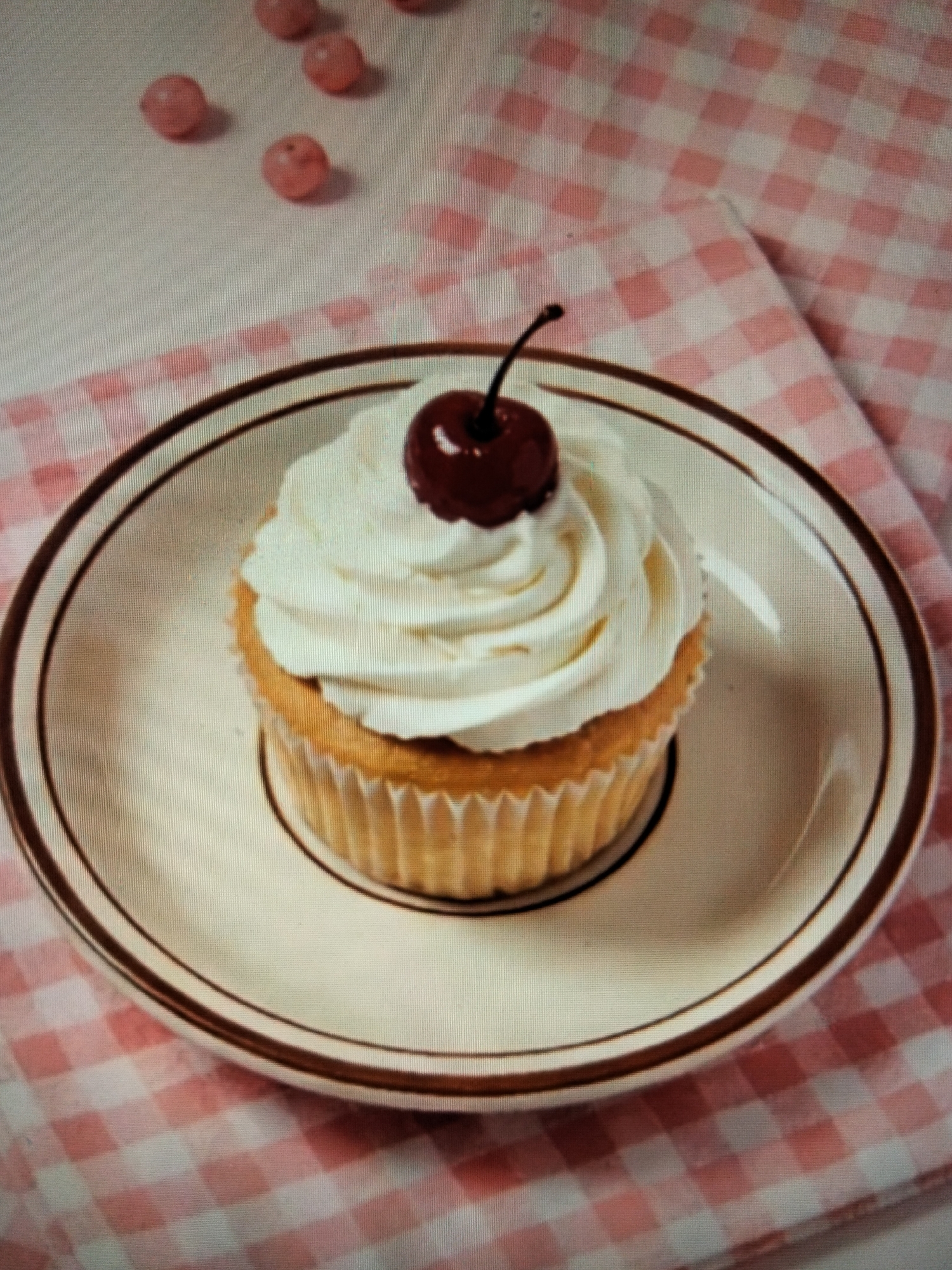 The image shows a single cupcake topped with white frosting and a cherry, presented on a light-colored plate with a dark rim. The plate is situated on a pink and white checkered tablecloth. In the upper left corner of the image, there are several small, round, pink objects scattered on the tablecloth, which appear to be candies or decorative beads. The background is largely out of focus, with the checkered pattern of the tablecloth being the most prominent feature. The scene is static and lacks any human presence or activity. The provided location information, "Payagaboh, Indonesia," does not appear to be represented or suggested by any elements within the image itself.