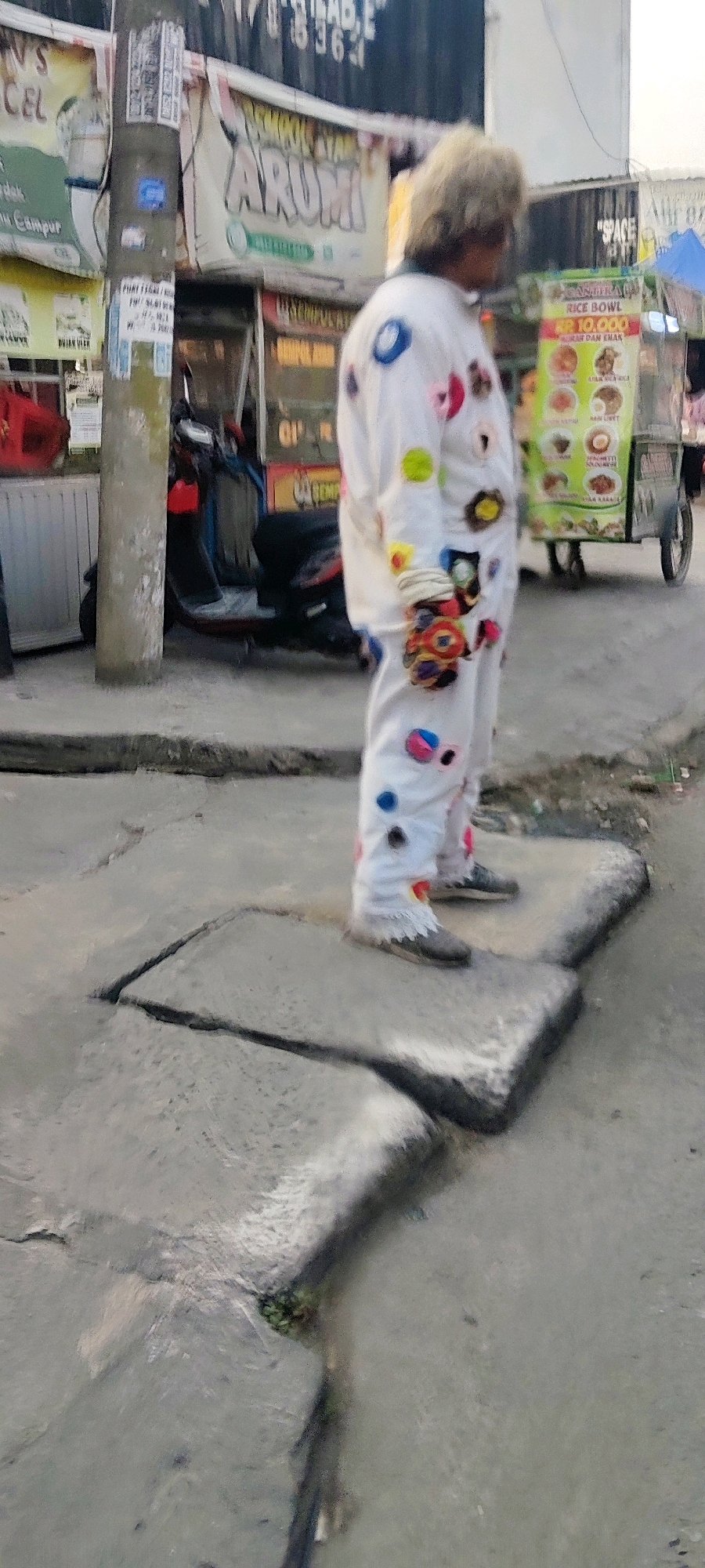 A person wearing a white jumpsuit decorated with colorful circles stands on a cracked concrete sidewalk in Mekarmukti, Indonesia. The jumpsuit has blue, pink, green, yellow, and red circles of various sizes. The person's hair is light-colored and appears fluffy. Their hands are gloved or bandaged with colorful fabric. In the background, there are food stalls with various signs, including one advertising "RICE BOWL." A motorcycle is parked behind the person. The environment suggests a street or market area with overhead banners and signage.