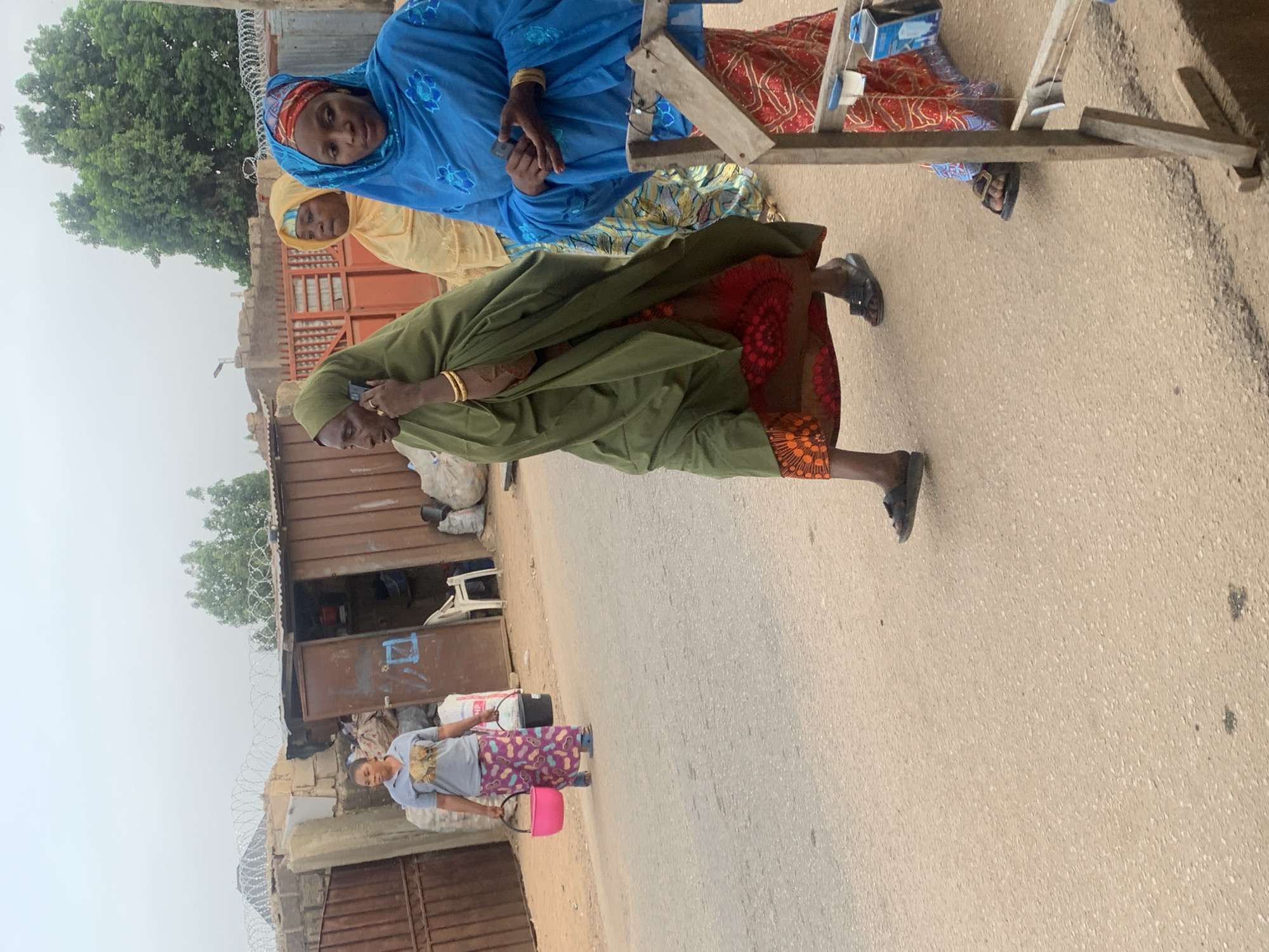 Three women in traditional attire stand together on a street. One wears a blue dress with gold jewelry and holds a cell phone. Another wears a yellow dress. The third woman wears a green robe over a patterned dress. A fourth woman walks down the street carrying a bucket. A building with a corrugated metal wall stands on the left side of the road. The location is likely Jos, Nigeria.