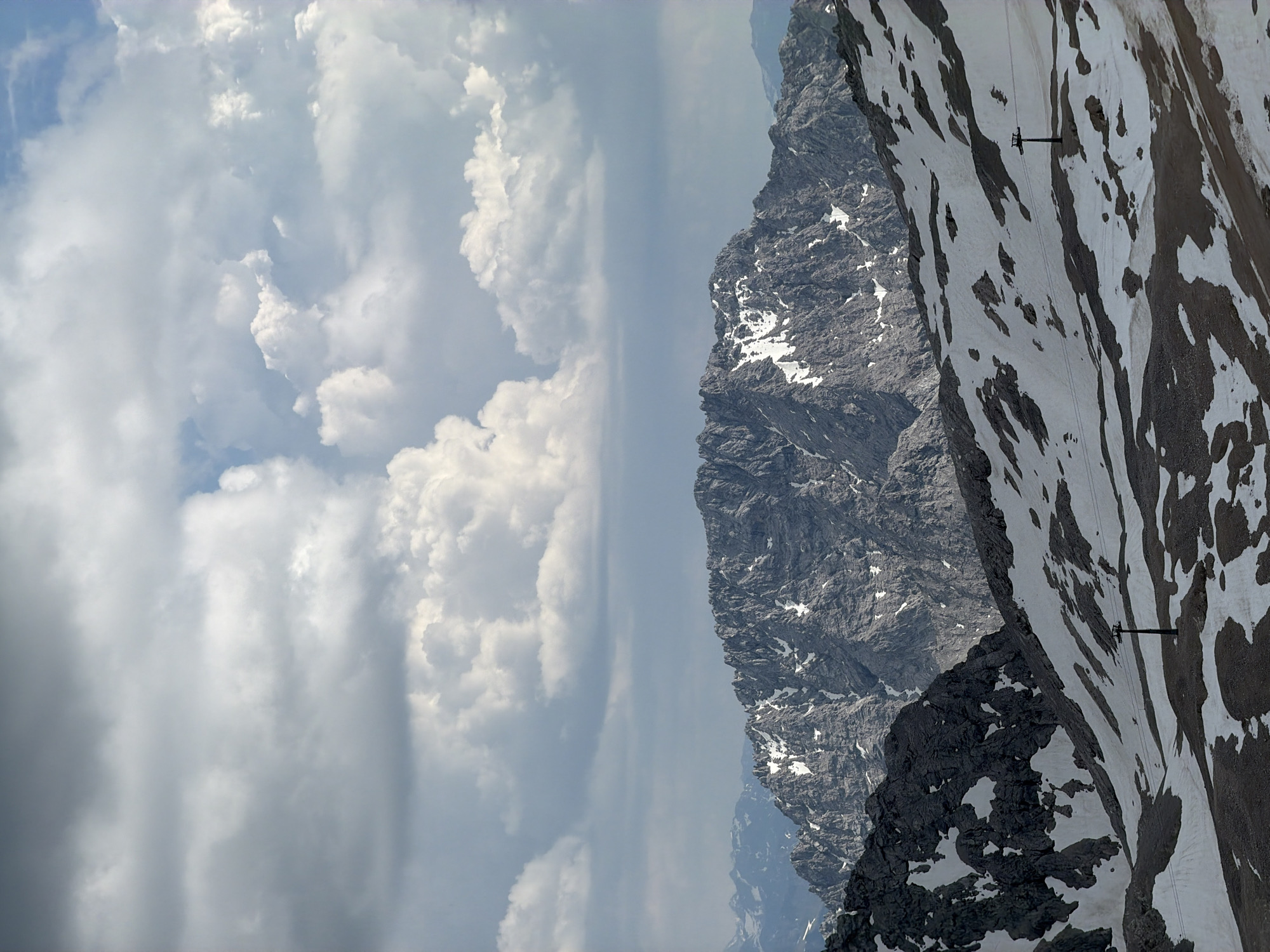 This image depicts a mountainous landscape with patches of snow and rock. In the foreground, a ski lift pylon stands on a snow-covered slope. The background features rugged, gray mountains with streaks of snow. The sky above is filled with dramatic, heavy clouds, ranging from dark gray to bright white. The scene appears to be taken from a high vantage point, offering a panoramic view of the alpine terrain. This is likely the Zugspitze area near Garmisch-Partenkirchen, Germany, given the characteristic jagged peaks and the presence of snow year-round. No people are visible in the image.