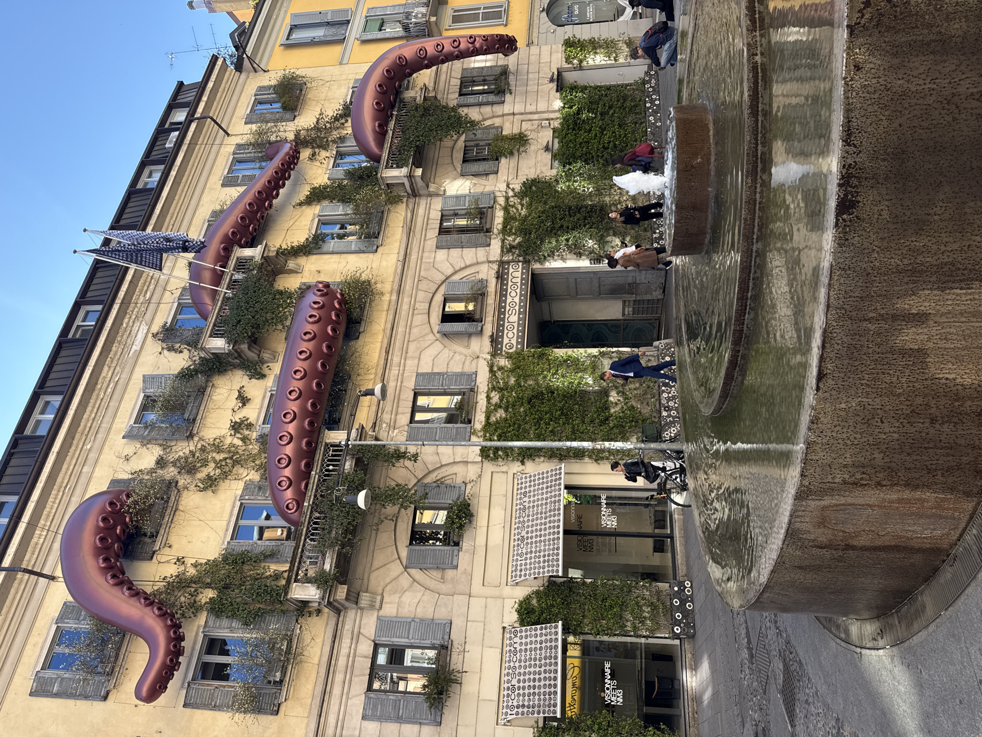 This image captures a vibrant street scene in Milan, Italy, featuring a multi-story building adorned with unique art installations and an active fountain in the foreground. The building, with a light yellow/beige facade, displays numerous windows fitted with grey louvered shutters. Patches of green ivy and climbing plants grow across the facade, spilling from balconies and around windows.
The most striking objects are several large, reddish-brown, sculpted octopus tentacles that dramatically emerge from and wrap around the building's structure, suggesting a temporary public art display. Each tentacle is detailed with visible suckers. On an upper balcony to the left, two dark blue flags with white patterns hang from a pole.
At street level, below the octopus tentacles, are shopfronts with large glass windows. Prominently visible text on these windows reads "VISIONNAIRE MEETS NM3," indicating businesses or an exhibition. Another sign further down reads "10-corso.como," a well-known concept store in Milan, confirming the location.
To the right, a large, rustic-colored circular fountain takes up a significant portion of the foreground, with water actively cascading over its edges and creating reflections. People are scattered throughout the scene; some stand near the fountain, observing, while others walk or cycle on the paved street. One person in a blue suit is visible near the fountain and building entrance, and another rides a bicycle.
The scene takes place on a clear and sunny day, indicated by the bright blue sky and well-defined shadows. The overall atmosphere is lively, showcasing an urban setting with an intriguing blend of architecture, nature, and contemporary art.