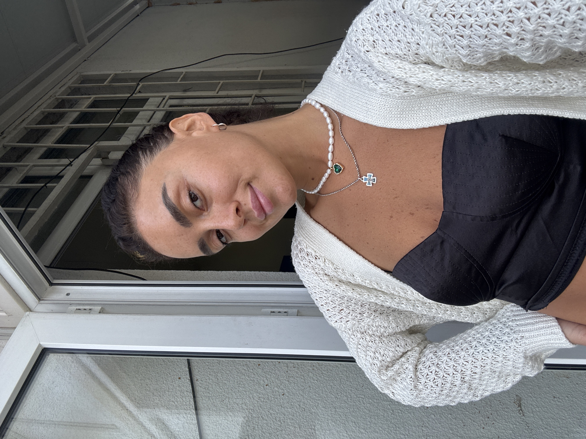 A woman with dark hair pulled back, freckles, and full lips poses for a selfie outdoors. She is wearing a white knitted cardigan over a black corset-like top. Two necklaces adorn her neck: one with pearls and a green heart pendant, and another with a silver cross pendant. She has a silver hoop earring in her right ear. The background suggests a residential area in Tbilisi, Georgia, with a white window frame, a barred window, and textured walls visible. The lighting indicates it is daytime.