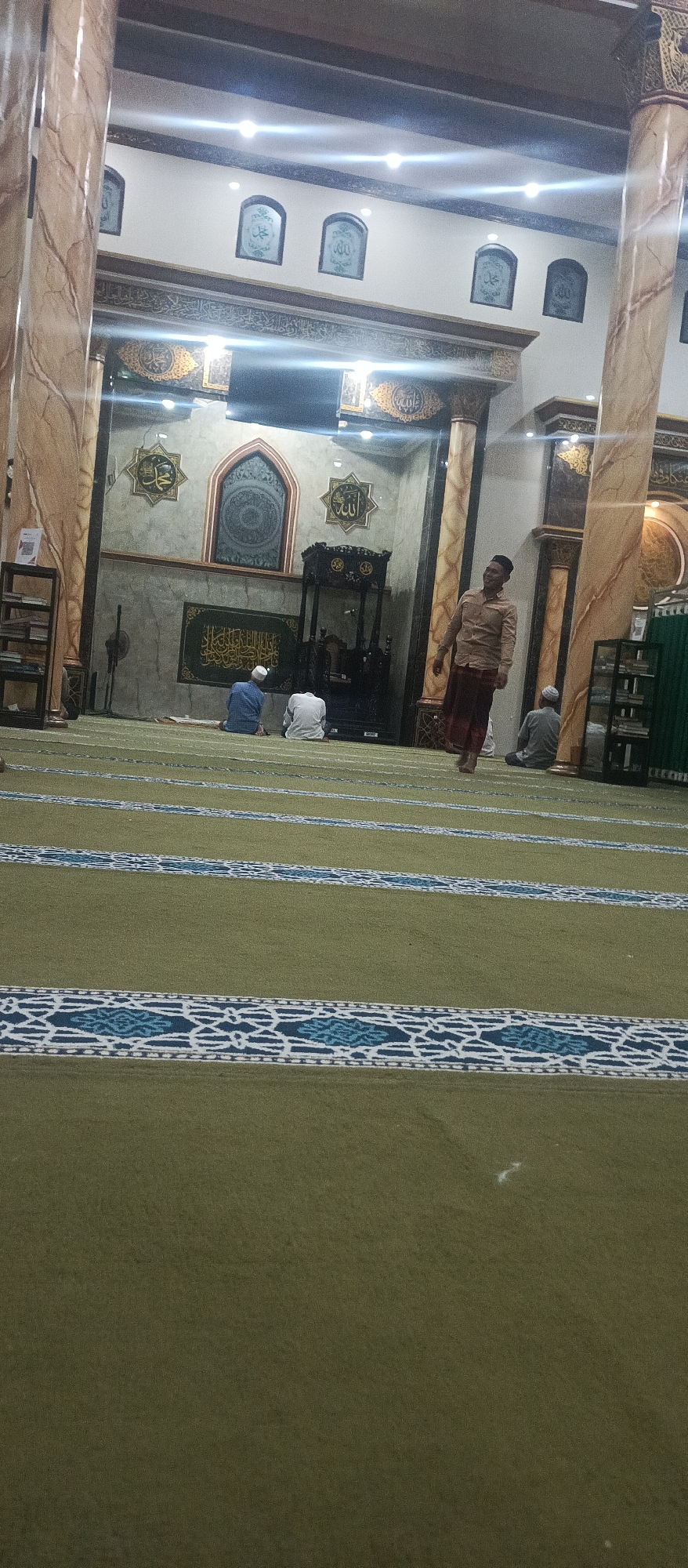 The image depicts the interior of a mosque in Arjasa, Indonesia. The central focus is the prayer hall, characterized by its green carpet with decorative blue and white stripes. Several individuals are present. In the foreground, a man is walking across the carpet, wearing a light-colored, long-sleeved shirt and a sarong. Further into the mosque, two men are seated on the carpet, facing forward, possibly in prayer or contemplation. A third man is also seated further back.
The mosque's architecture features tall, marble-patterned columns. The walls are adorned with Islamic calligraphy and decorative motifs. Above the prayer area, there is a pulpit and an ornate archway. The lighting appears to be a mix of overhead lights and ambient illumination. The scene suggests a calm and reverent atmosphere within the mosque.