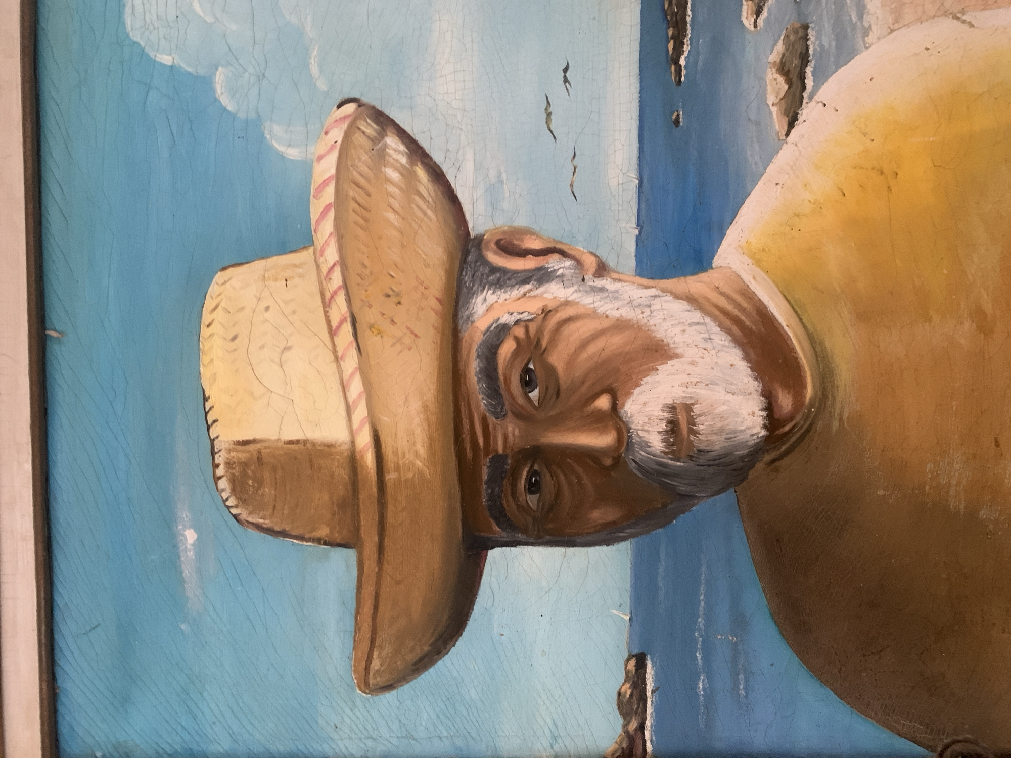 The image depicts a painting of an elderly man wearing a straw hat. The background features a blue sky and a body of water with rocky outcroppings. The man has a white beard and mustache. The painting appears to be somewhat aged, with visible cracking in the paint. No explicit details link the painting to the city of Santa Ana, Venezuela.