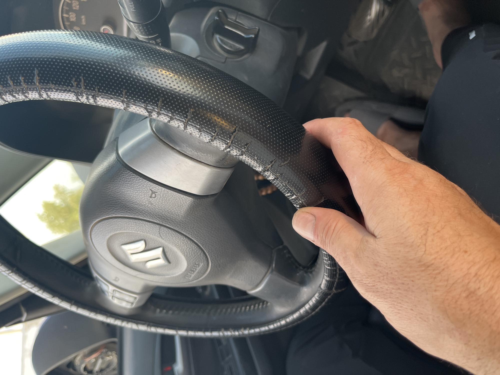 A person's left hand grips a worn steering wheel inside a vehicle. The steering wheel is black with a textured grip, and the center displays the Suzuki logo. The dashboard and part of the driver's seat are visible. The image is taken from a low angle, looking up towards the steering wheel. The background shows a blurred view of trees and sky. The overall scene suggests the interior of a car, likely in a parking lot or on a street. No specific indicators of the city of Шумейка, Russia are present in the image.
