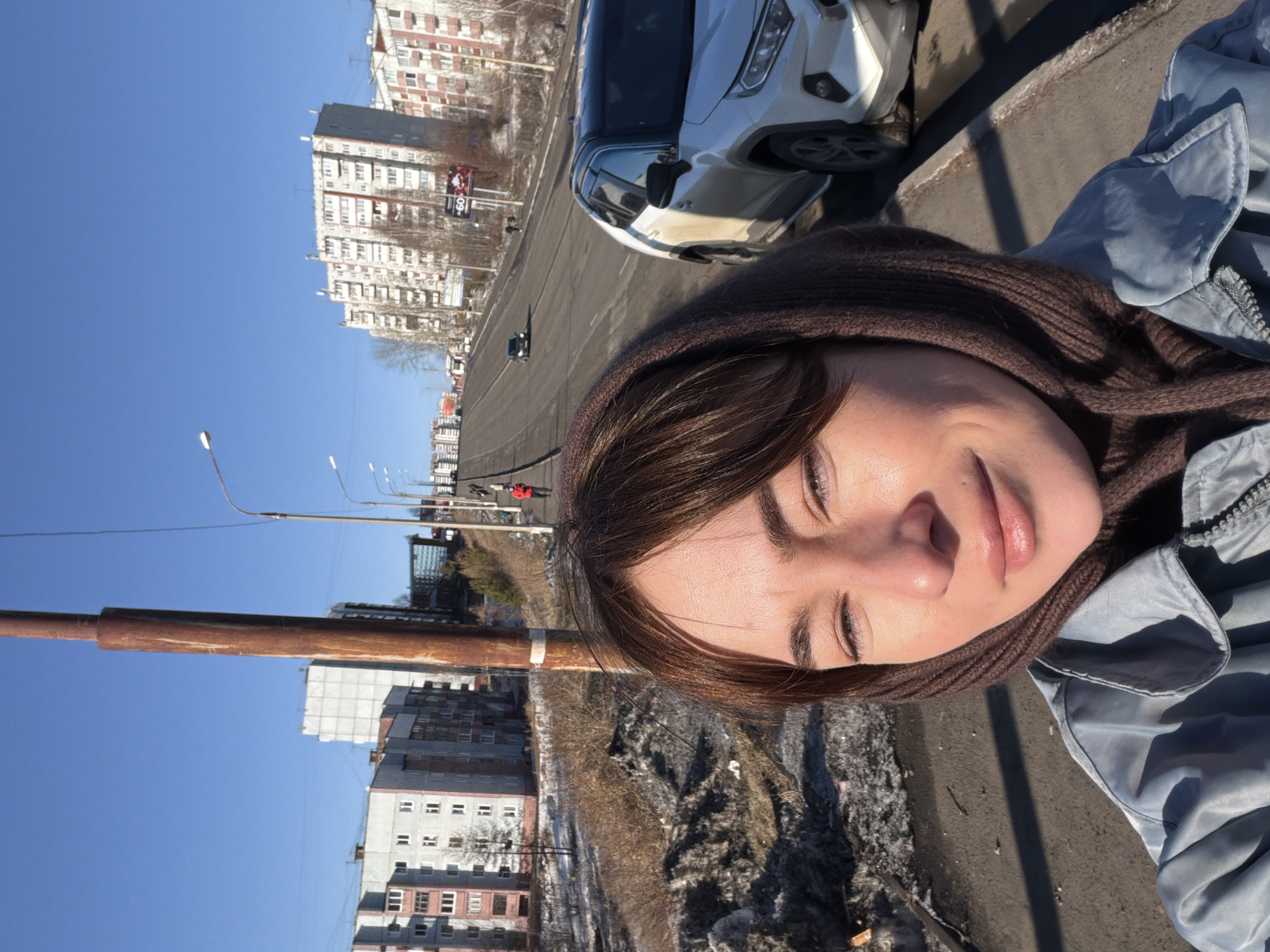 A slightly rotated medium shot of a woman standing outside on a clear, sunny day near a road in Ust-Ilimsk, Russia. She is wearing a blue jacket and a brown scarf/hood. In the background, there are several buildings typical of a Soviet-era cityscape, a parked white car, a road with some vehicles, a rusty lamppost, and patches of melting snow on the ground. The sky is clear and blue. The woman is looking at the camera with a slight smile.