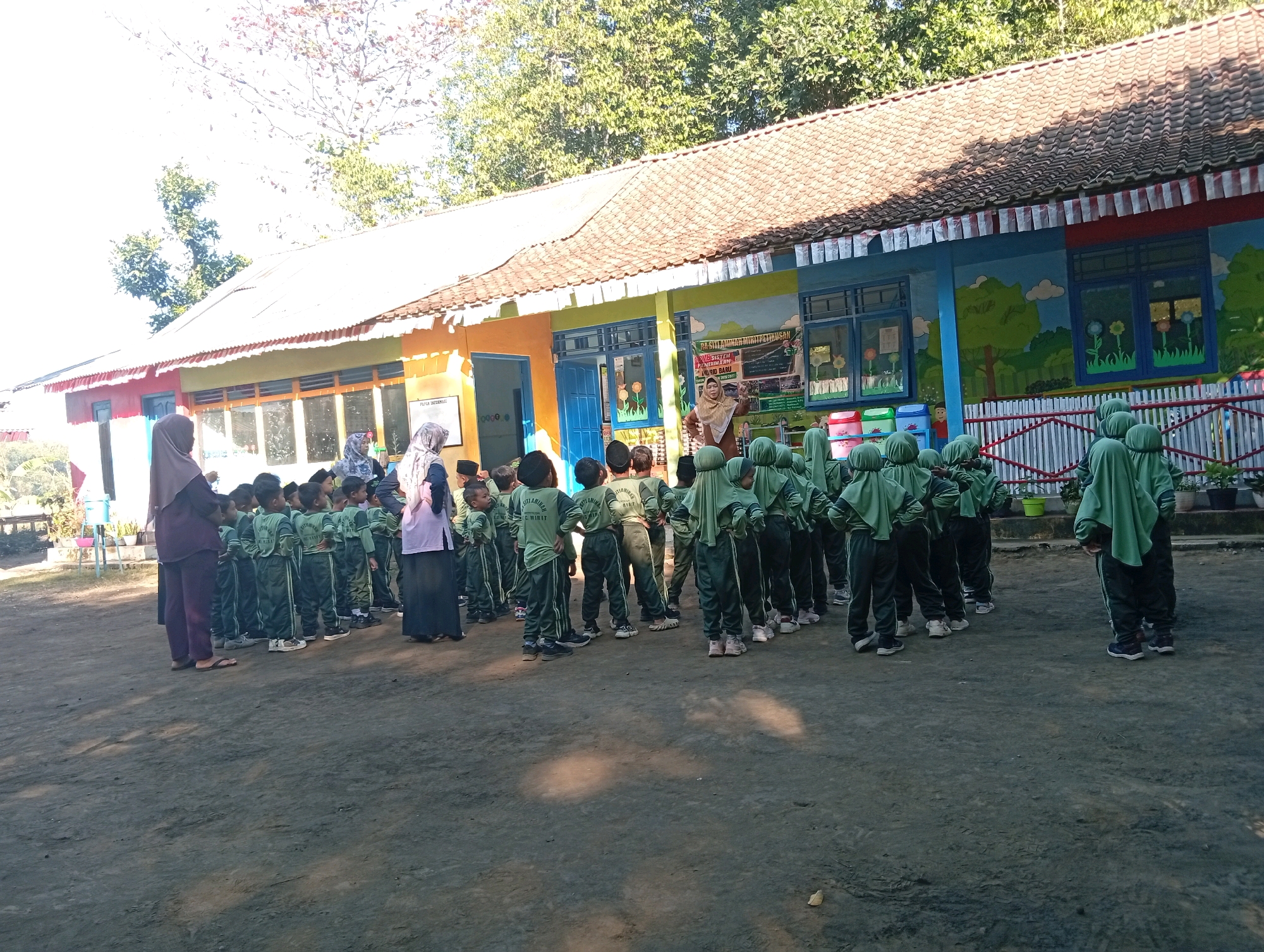 A group of young children, likely students at a school, are gathered outdoors in what appears to be a schoolyard. The children are all dressed in matching green uniforms, consisting of long-sleeved shirts and trousers, and some are wearing headscarves. They are standing in neat rows, facing towards a building.
The building behind them is a single-story structure with a tiled roof. It has a colorful facade, painted in shades of orange, yellow, green, and blue, with murals depicting trees and clouds. There are several windows with blue frames and some doors. The building appears to be a school or educational facility.
In front of the building, there are several adults, possibly teachers, interacting with the children. One adult, wearing a purple headscarf and matching outfit, stands to the left of the frame, appearing to supervise the children. Another adult, wearing a light brown headscarf and a light brown top and dark trousers, is standing and gesturing towards the children.
The ground in front of the building is a dirt or gravel surface, with some scattered debris. The area is bathed in sunlight, casting shadows on the ground. In the background, there are trees, indicating a natural or semi-rural environment. The image likely depicts a school event or a regular school day activity at a school in Miritpetikusan, Indonesia.