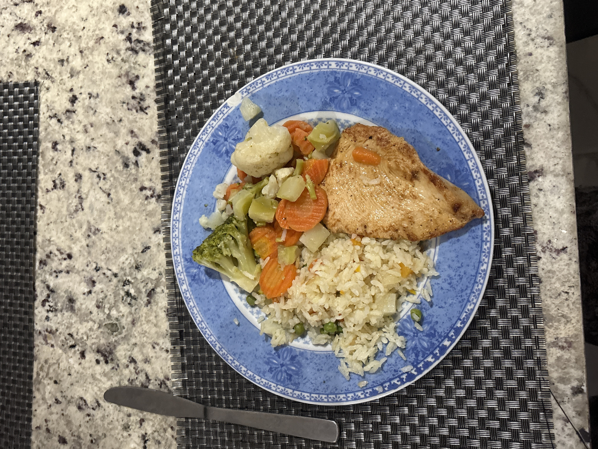 The image shows a plate of food on a table. The plate is blue and white and contains grilled chicken breast, rice with peas, broccoli, cauliflower, and carrots. A knife is placed on the left side of the plate. The table has a patterned placemat. Part of the table is a granite countertop. The image location cannot be determined to be San Bernardino Tlaxcalancingo, Mexico based on the visual information.